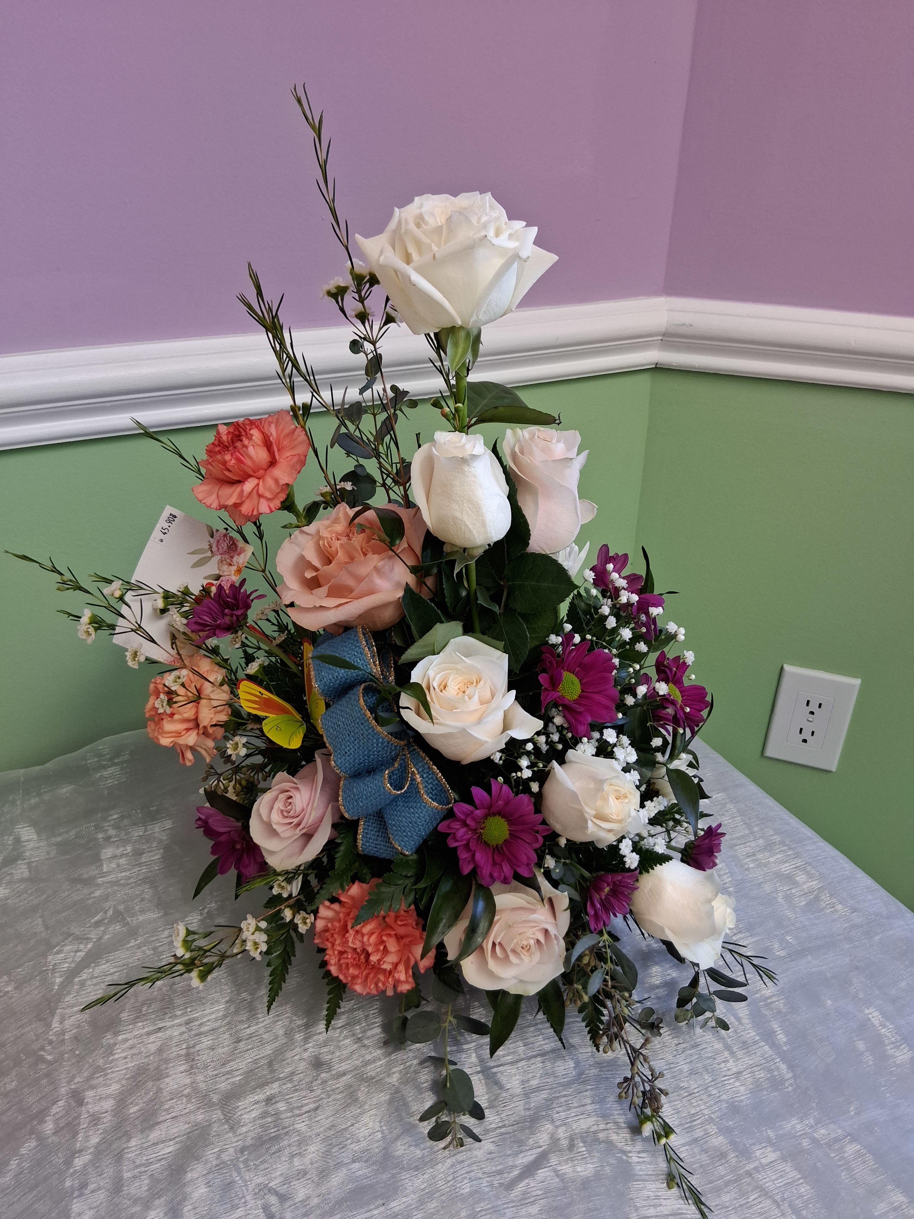 Elegant Arrangement for every occasion   - Soft blush and white roses meet coral carnations and purple daisy mums in a garden-style flower arrangement made for celebrations and sentimental moments alike - perfect as a birthday bouquet, anniversary surprise, or just-because gift. The layered, airy composition and playful butterfly pick give the recipient a feeling of being truly seen: romantic, fresh, and thoughtfully handcrafted.  This arrangement mixes roses, carnations, daisies, and delicate white filler flowers arranged at varying heights for a full, natural silhouette. Colors include white, pink, purple, and orange, tied with a blue burlap ribbon and finished with a paper tag for your message. The loose, feminine styling offers contrast and movement, so it reads beautifully on a mantel, dining table, or bedside.  Choose this arrangement when you want to make someone feel celebrated and cherished - whether for a milestone or a quiet act of kindness. Available in a standard, ready-to-gift size; for best life, keep flowers in fresh water, trim stems slightly, and display away from direct heat and drafts.