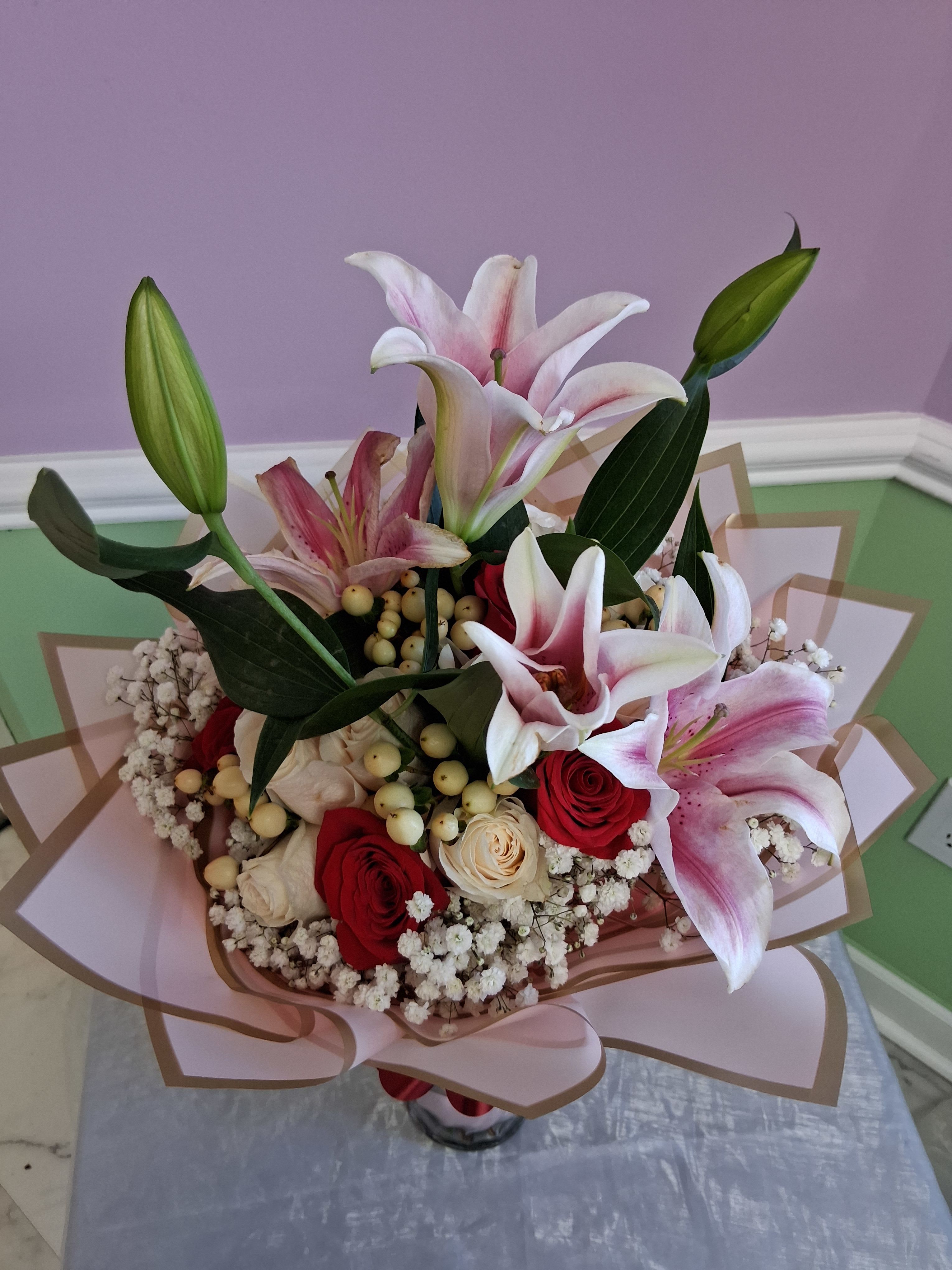 One dozen Red and White Roses with Lilys - Large pink-and-white lilies soar above a tight cluster of red and cream roses in this romantic flower arrangement-perfect as an anniversary bouquet, birthday surprise, or any gift celebrating love and romance. The dramatic height of the lilies paired with the classic dozen roses creates an immediate "wow" that makes the person you're thinking of feel truly seen and cherished.  The arrangement mixes lush lilies, red and cream roses, and small white filler blooms with pale, berry-like stems for texture, all arranged in a balanced, layered silhouette. Soft blush-and-tan decorative wrapping paper frames the stems and the whole design comes presented in a clear glass vase, offering both polish and practicality. Colors include shades of pink, red, white, and green for a timeless, feminine palette.  This is the kind of bouquet that reads as both celebratory and intimate-ideal for anniversaries, romantic gestures, milestone birthdays, or simply saying "I love you." Arrives vase-ready for easy display; remove any wilted petals or spent blooms and refresh the water every 2-3 days to keep the lilies and roses looking their best.