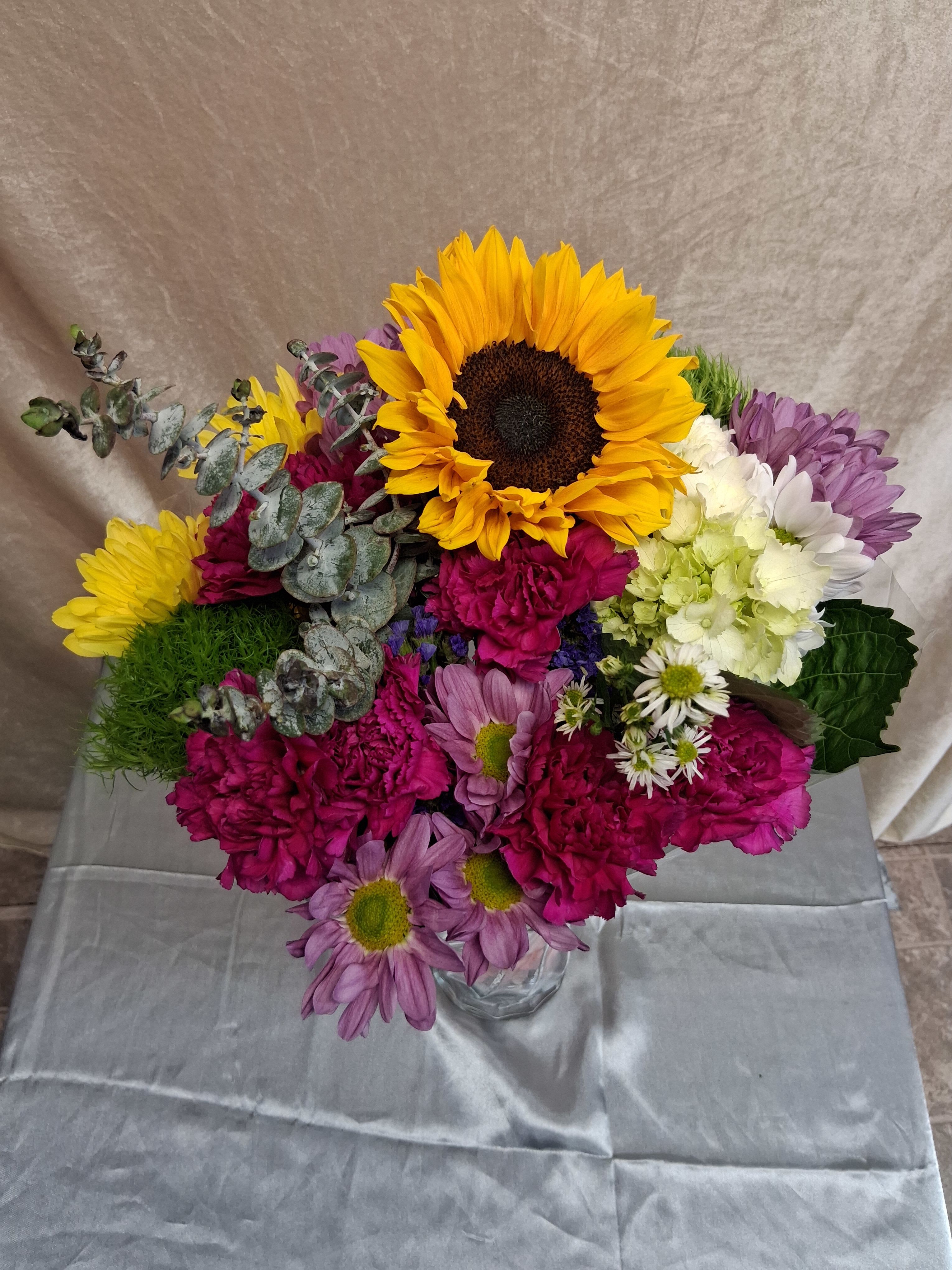 Wild mix bouquet  - A bold golden sunflower steals the show in this wild mix bouquet, a cheerful flower arrangement perfect for birthdays, congratulations, or a just-because surprise. The bright, sunny center paired with pink, purple, yellow, and white blooms creates an immediate feel-good impact-she'll see the colors and feel genuinely celebrated the moment the vase is set down.  This bouquet features a large sunflower surrounded by rounded chrysanthemum heads and daisy-like flowers in magenta, lavender, white, and pale green, with silvery eucalyptus-style foliage adding cool contrast and texture. Arranged in a simple clear glass vase, the dense, colorful cluster reads fresh and seasonal, letting the saturated petals take center stage while the glass keeps the look modern and unfussy.  Ideal for a friend, parent, or coworker who loves vibrant, textured blooms, this arrangement sends a bright, uplifting message without being over the top. Vase included; to keep the bouquet looking its best, refresh the water every few days and trim stems at an angle when you change the water.