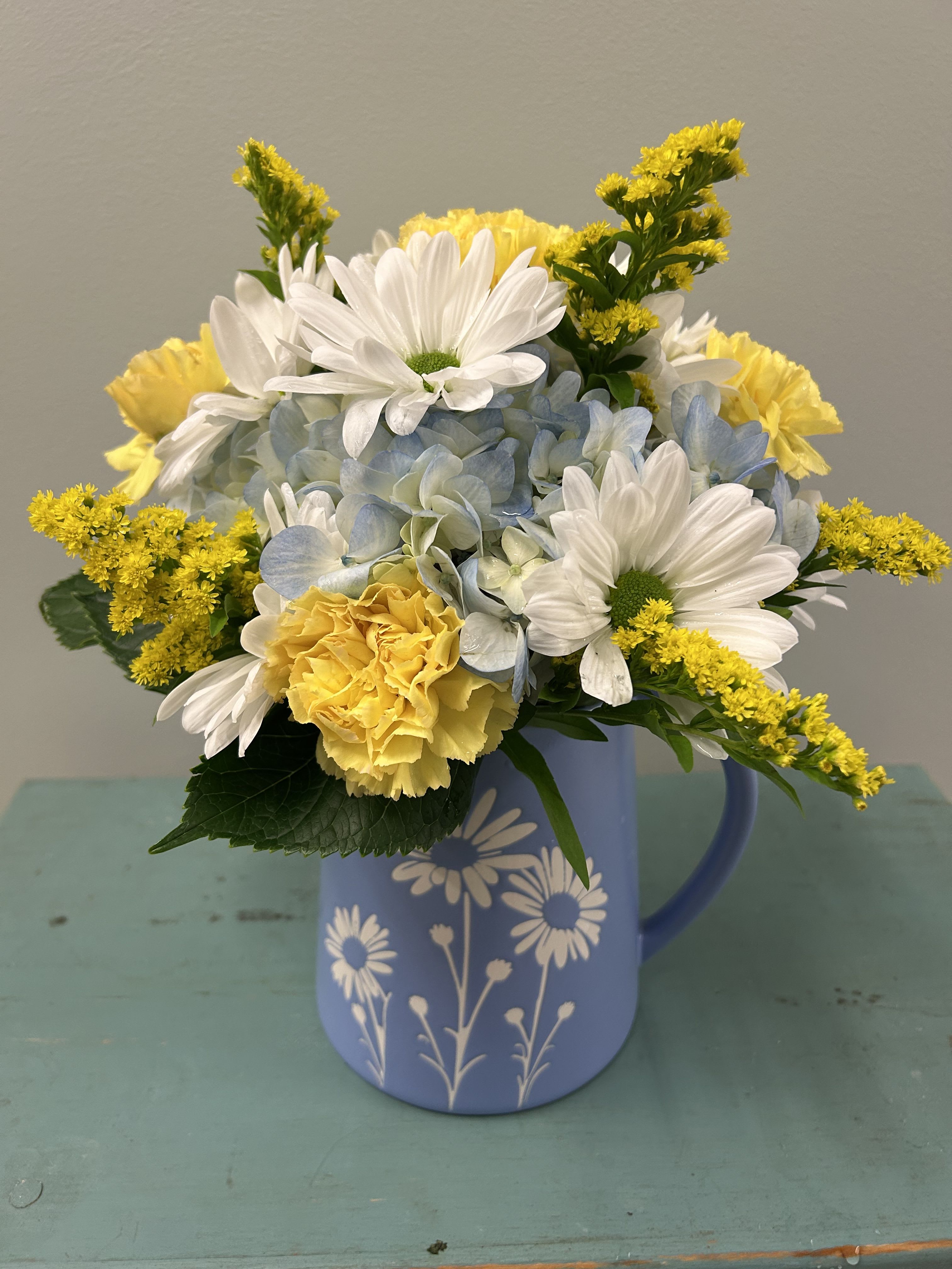 Daisy Pitcher - Bright, cheerful daisies and sunny yellow carnations mingle with soft blue hydrangea blooms in this Daisy Pitcher - a casual flower arrangement perfect for birthdays, Mother's Day, get well wishes, or simply brightening someone's day. The mix of white daisies, crisp yellow accents, and pale blue creates an uplifting, fresh mood that makes the recipient feel seen and cared for, whether you're saying "happy birthday," "thinking of you," or "thank you."  Arranged in a light blue ceramic pitcher decorated with a white daisy motif, the flowers are gathered into a compact, rounded dome that reads full and abundant on tabletops. The design pairs large open faces (daisies and hydrangea clusters) with smaller textural filler blooms for contrast and visual depth. The pitcher serves as both vase and keepsake, offering an easy, ready-to-display gift that brings a casual, homey charm to kitchens, living rooms, or bedside tables.  This size fits well as an everyday centerpiece or a small, heartfelt gift for the person you want to lift up-no vase hunting required. For best enjoyment, keep the stems in fresh water and trim them slightly if needed to maintain the dome shape; replace water regularly to extend the life of the blooms.