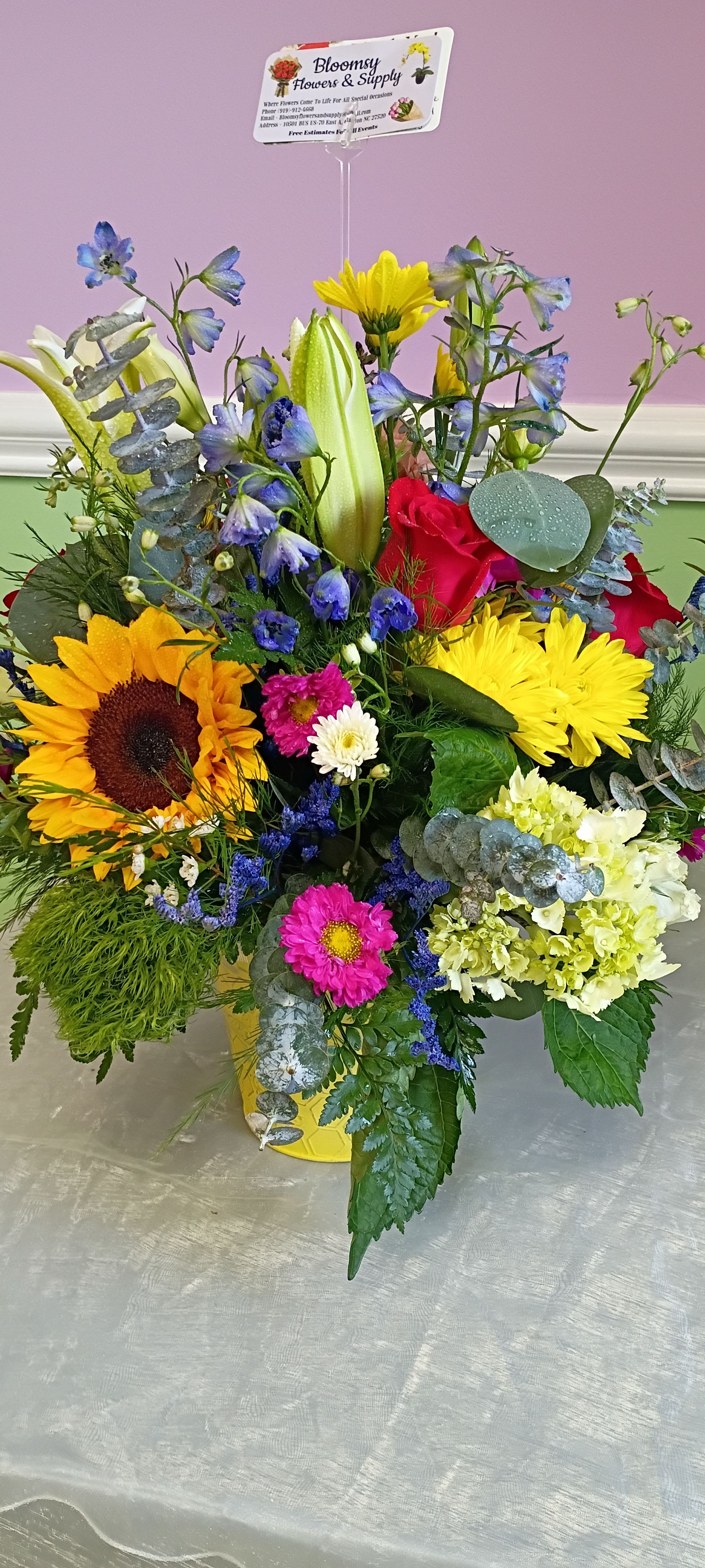 Wildflower glass vase arrangement  - Bold yellow sunflowers and golden chrysanthemums set a sunny, celebratory tone in this wildflower glass vase arrangement, an ideal birthday bouquet or congratulations gift that reads joyful from across the room. Pink roses and white lilies soften the palette while sky-blue delphinium-like blooms and daisies add playful contrast, so the person you're thinking of will feel seen and uplifted by its cheerful, garden-fresh energy.  This large, mixed bouquet is arranged full and layered for a lively, abundant look: dense focal flowers sit at the center, airy stems and eucalyptus provide height and texture, and varied bloom shapes keep the eye moving. The arrangement is presented in a bright yellow container with a clear plastic sign holder tucked into the design for a personal note or small message.  Perfect for birthdays, new jobs, housewarmings, or any celebration where you want to make someone feel truly noticed, this piece arrives ready to display and brighten a room. Care: keep stems in fresh water, trim stems every few days, and place away from direct heat to prolong bloom life.