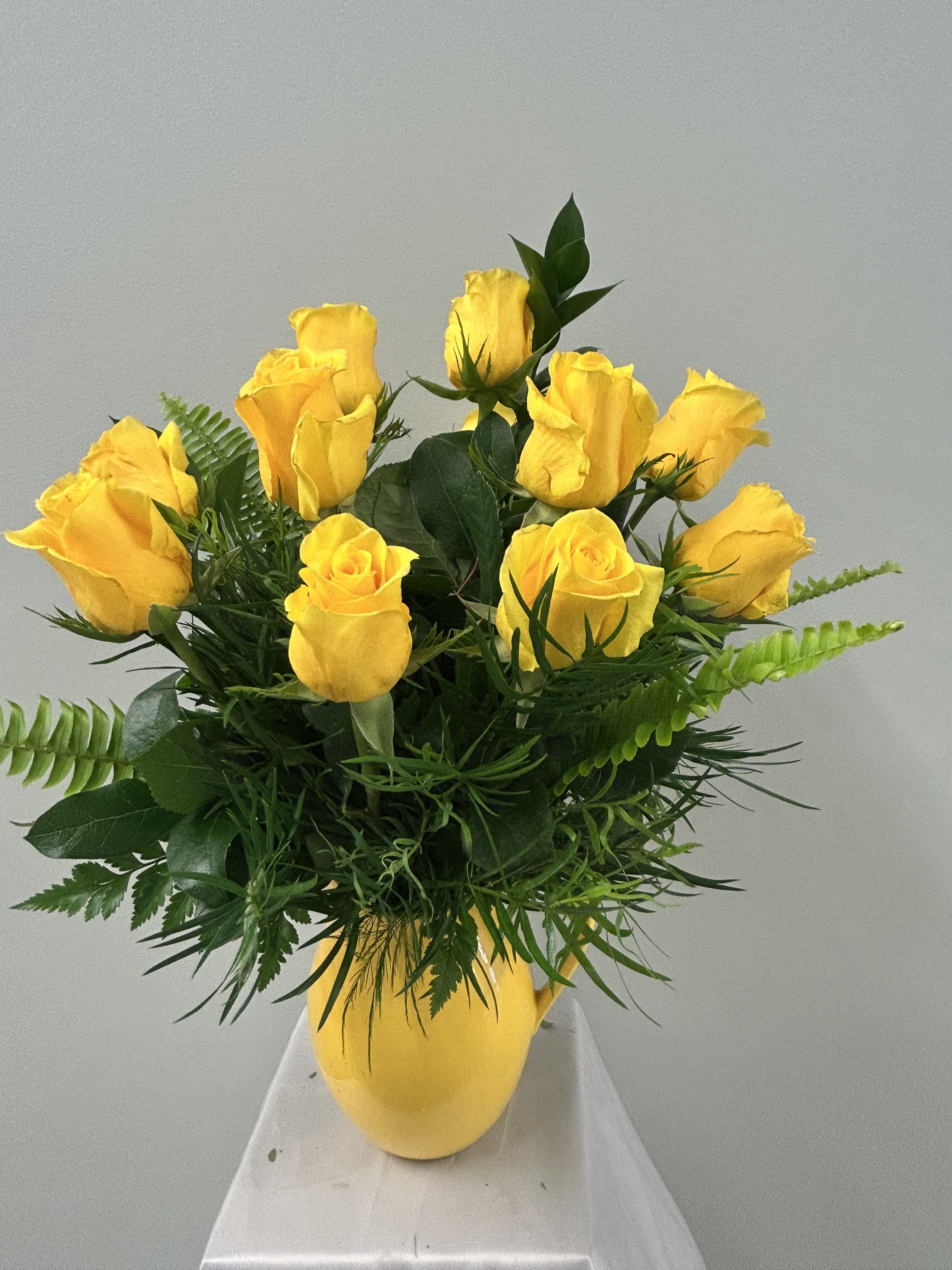 Pitcher of Sunshine - Sunshine in a pitcher-bright yellow roses rise above a collar of mixed green foliage in a smooth yellow ceramic pitcher, making this flower arrangement an instant mood-lifter for Mother's Day, birthdays, anniversaries, or a cheerful get-well surprise. The vivid yellow and deep green contrast gives a fresh, energetic feel that reads as warm gratitude, friendly celebration, or simple "thinking of you" joy for housewarmings, retirements, Father's Day, and casual occasions.  The arrangement includes a cluster of a dozen yellow roses surrounded by lush mixed foliage, arranged low-to-medium in a yellow ceramic pitcher that doubles as a keepsake vase. The compact, handheld size fits tabletops and counters-ideal for Spring and Summer décor or to brighten an office for Admin Professionals' Week, Boss's Day, or a colleague's congratulations. Care is simple: keep water fresh and trim stems every few days to extend bloom life and keep the display looking its sunny best.