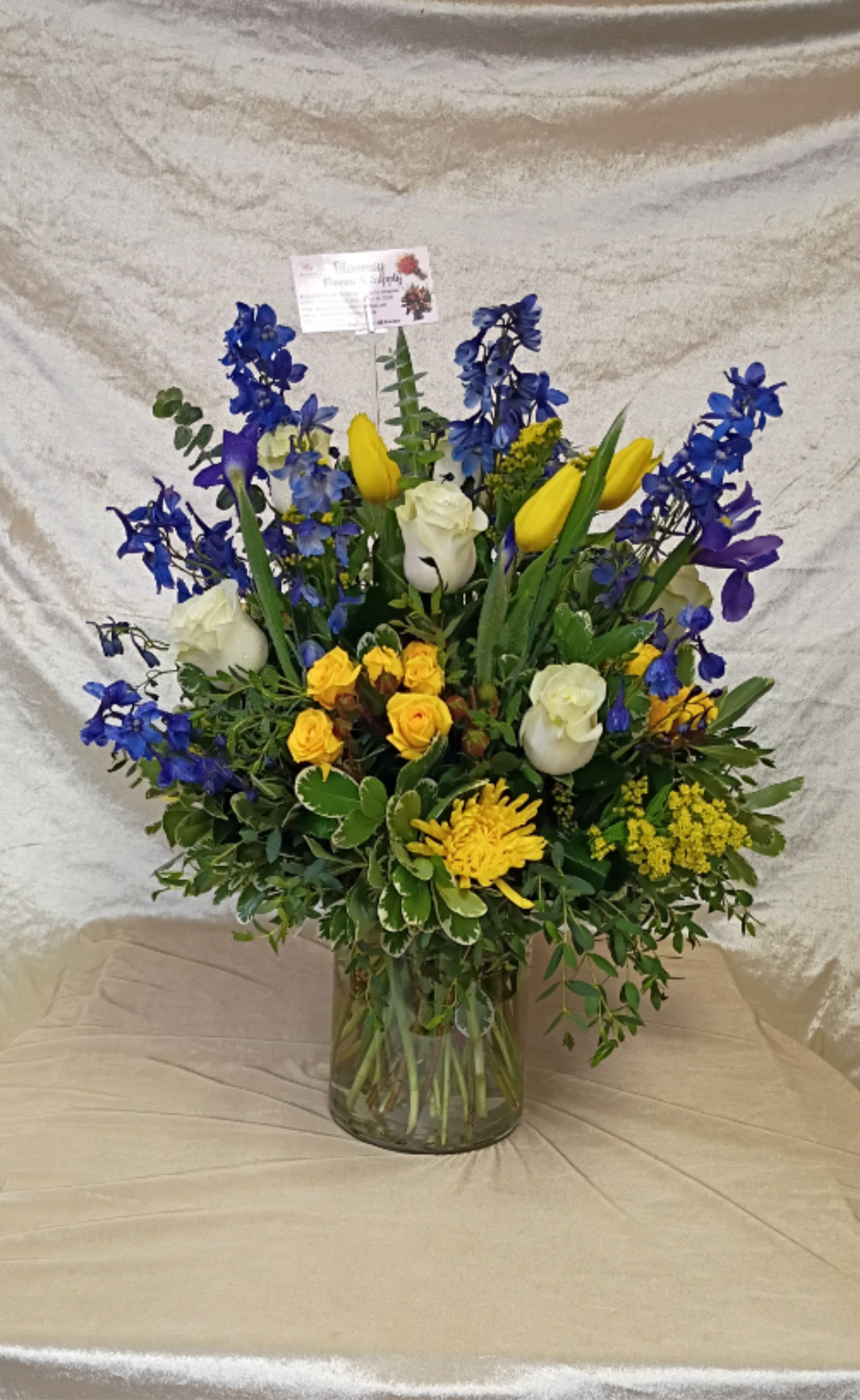 Elegant arrangement with roses and tulips  - Tall blue delphinium spikes rise above sunny yellow tulips and creamy white roses in this cheerful flower arrangement, perfect as a birthday bouquet, congratulations gift, or everyday pick-me-up. The vivid blue-and-yellow palette creates a lively, celebratory impression while the white roses and daisylike accents soften the look so the recipient feels genuinely seen and uplifted.  Arranged upright in a clear glass vase, the design pairs varied textures-delphinium's vertical drama, the smooth tulip petals, clustered chrysanthemums and spray blooms-for a full, layered composition. Dense mixed greenery fills the base for a lush, garden-like feel; a discreet floral pick adds structure so stems hold their elegant shape. Ideal for brightening a home, office, or party, this arrangement arrives ready to display and brings long-lasting color and freshness to the moment.