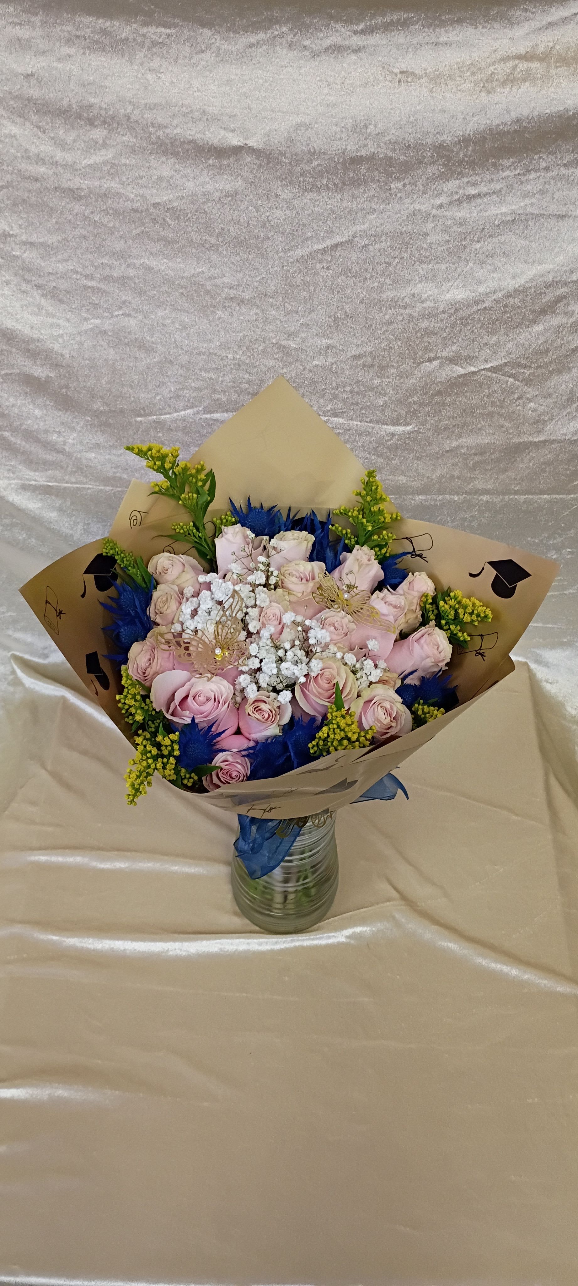 Two dozen pink roses for graduation style blue - Pale pink roses gather in a soft, romantic cluster centered on a cloud of white baby's breath-an elegant graduation bouquet that reads both celebratory and heartfelt. The two dozen pink roses make a strong visual statement for graduation or commencement gifts, while vivid blue accent flowers and a large sheer blue ribbon reinforce the school‑color or class pride theme so it feels tailored to the graduate.  This wrapped arrangement pairs the roses with small yellow filler sprays and touches of green for brightness and contrast, finished with a gold butterfly ornament for a whimsical flourish. The flowers arrive tucked in tan brown paper printed with graduation cap motifs and presented in a clear ribbed glass vase, making it ready to display. Dimensions and vase style are designed for easy placement on a desk or dining table.  Give this to the grad who deserves to feel celebrated-the palette says congratulations and the generous two dozen roses will make her feel truly seen. For care, keep the vase topped up with fresh water and trim stems slightly every few days to extend bloom life; the ready-to-display wrapping and vase make same-day gifting simple and polished.