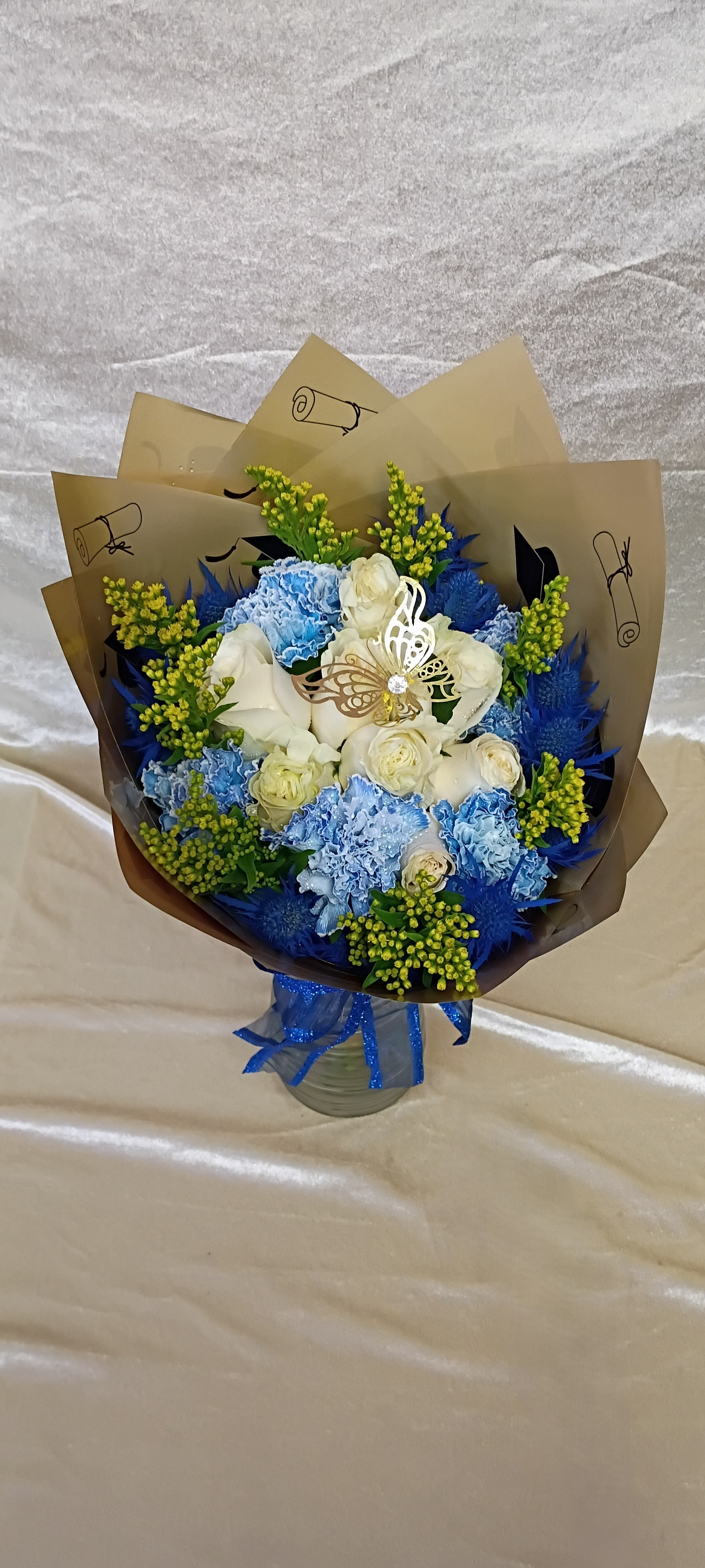 Graduation bouquets Female and Male Roses and Carnations - Crisp blue and white roses paired with pale blue carnations create a celebratory graduation bouquet that reads both classic and youthful-perfect as a graduation bouquet for her or a tasteful congratulatory gift for him. The cool-toned mix of vivid blue filler blooms and white roses is brightened by small yellow-green spray accents for contrast, so the person you're honoring will feel recognized and genuinely celebrated on their big day.  Wrapped in layered brown paper and tied with a shiny blue ribbon, the arrangement arrives gift-ready with a polished, slightly formal look. A smaller option is presented in a clear glass jar wrapped in tan paper printed with graduation caps and diplomas, finished with a glittery blue ribbon and butterfly ornament for a playful, school-ceremony theme. Sizes and vase options vary-choose the wrapped bouquet for a hand-held presentation or the jar style for a keepsake display-and expect long-lasting stems with simple care: fresh water, recut stems every few days, and a cool location away from direct sun.