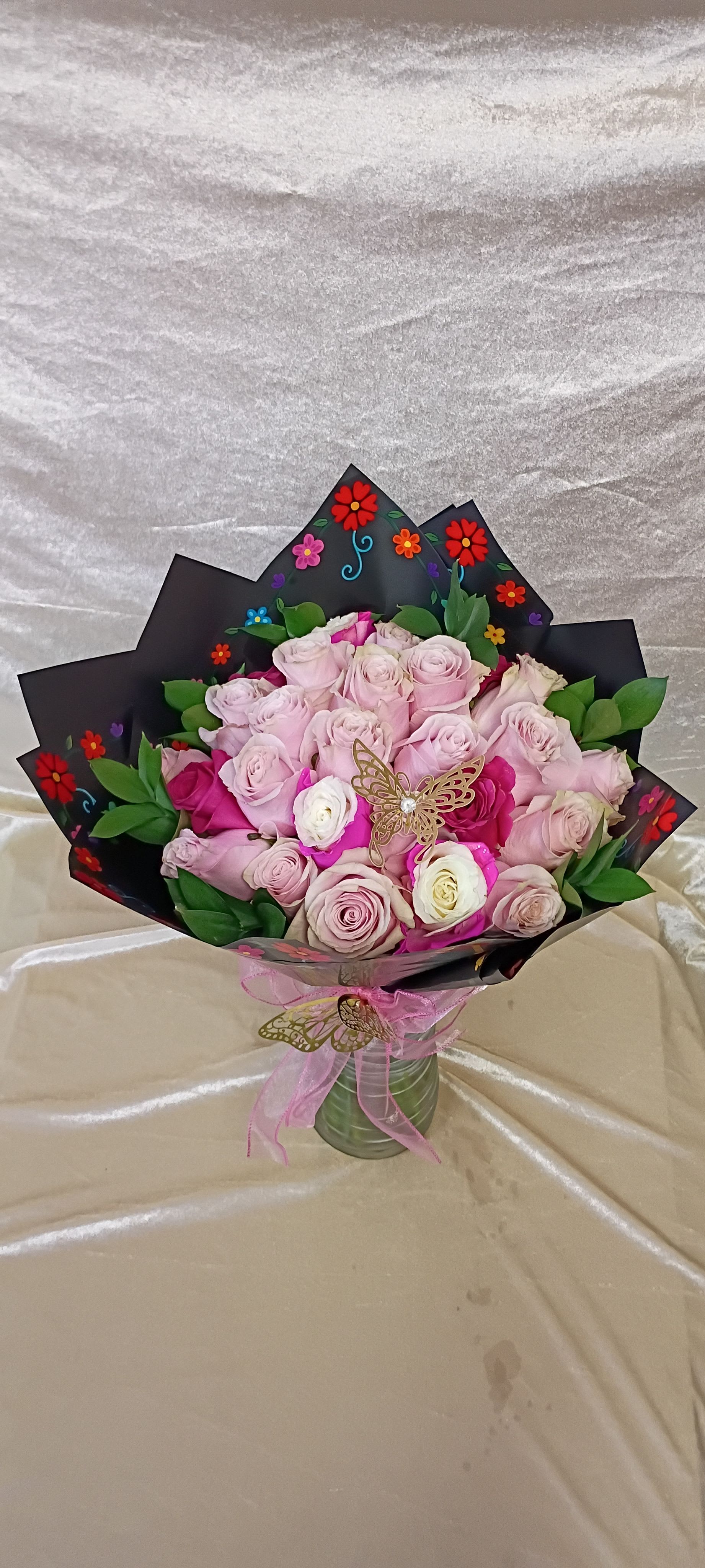Two dozen mix pink roses bouquet  - A tightly packed spray of two dozen pink roses creates an immediate romantic statement-perfect as a birthday bouquet, anniversary roses, or a Valentine's Day gift for the person you want to make feel cherished. Pale pink petals mingle with a few white blooms and deeper magenta accents at the center, giving the bouquet depth and a soft, scented presence that reads feminine and celebratory.  The roses arrive arranged in a clear glass vase and wrapped in a tall black decorative sleeve patterned with small colorful floral motifs, the dark wrap making the pastel blooms pop. A pink ribbon tied around the vase and a delicate butterfly ornament complete the presentation, lending a playful, gift-ready finish. The contrast of soft pinks, crisp white, and green foliage against the black decorative wrap makes this arrangement equally suited to romantic moments, thank-you gestures, or a joyful just-because surprise.  This is the kind of bouquet that will make her feel seen and celebrated-elegant on a mantel or ready to present at a table. Available in the pictured clear glass vase; standard care (fresh water and a cool spot out of direct sun) will keep the roses looking their best through the week.