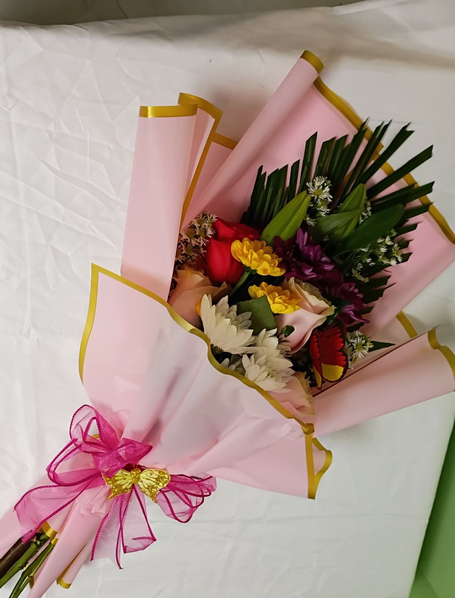 Presentation bouquet  - A jubilant hand-tied presentation bouquet that reads like a celebration in bloom - perfect as a birthday bouquet, congratulations gift, or a romantic surprise. The mix of roses, chrysanthemums, daisies, lilies and seasonal fillers creates an immediate, cheerful impact in pink, white, yellow and mixed jewel tones so the person you're thinking of will feel noticed and celebrated the moment they see it.  Tightly arranged for a full, rounded silhouette, the flowers are wrapped in soft pink paper with elegant gold trim and finished with a sheer pink ribbon bow with a small gold accent for a polished, gift-ready look. Colors range from soft pastels to bright pops of red, purple and yellow to keep the arrangement lively and texturally interesting - velvety rose petals, crisp daisy centers, and the sculptural forms of lilies and chrysanthemums all play together.  This bouquet is made to arrive ready to display and delight: ideal for sending congratulations, a heartfelt birthday wish, or to say "I'm thinking of you" in a way that feels celebratory and feminine. For best freshness, trim stems and change the water daily when you place it in a vase; orders are hand-tied and designed to make an immediate impression on the recipient.