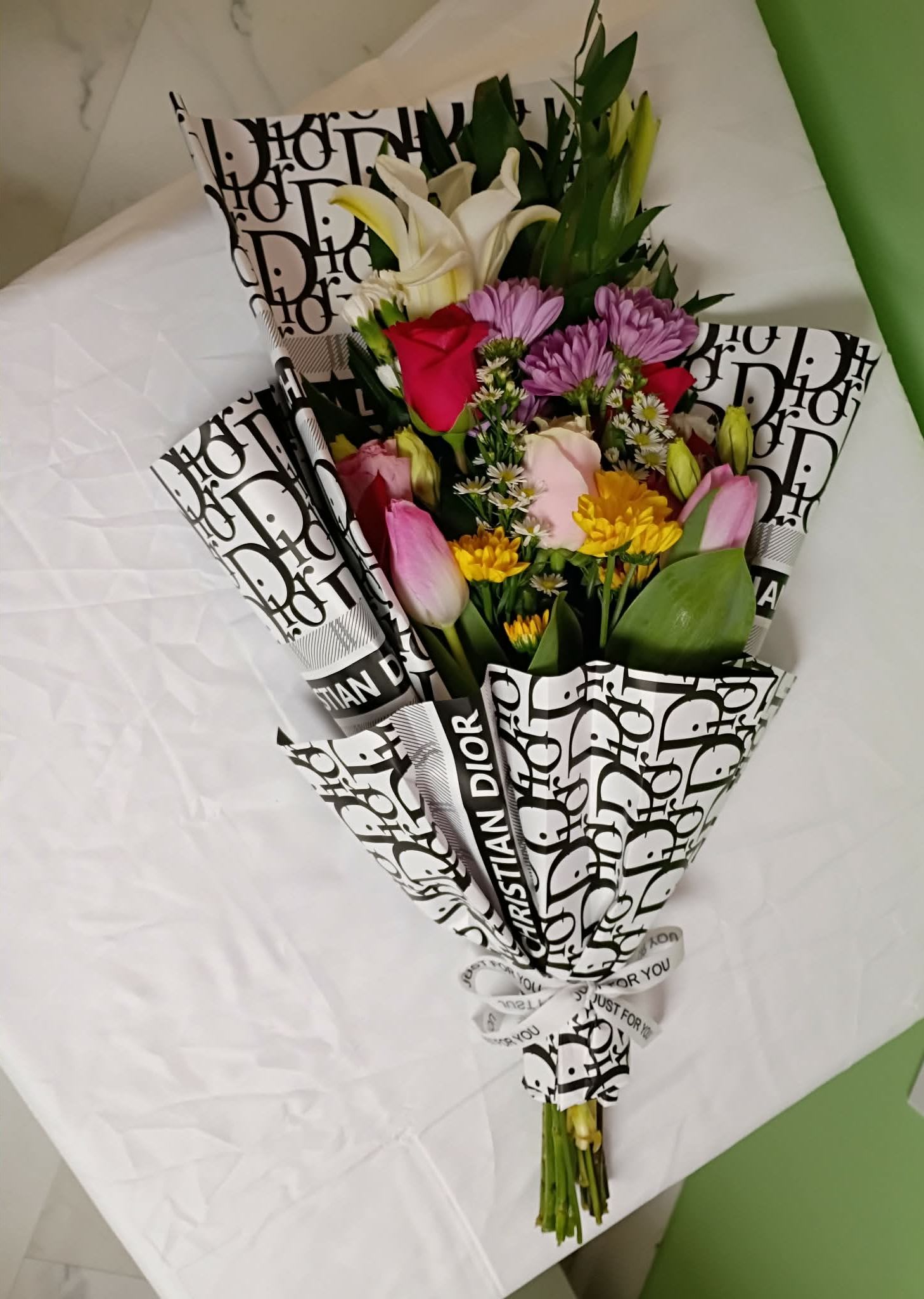 Presentation Bouquet mix flower - Bright pink roses and trumpet-shaped white lilies take center stage in this presentation bouquet, a cheerful gift-ready arrangement perfect for birthdays, anniversaries, or a thoughtful thank-you. The mix of pink tulips, purple daisy-like blooms, and little yellow accent flowers creates a lively, celebratory palette that will make the person you're thinking of feel genuinely seen and celebrated.  Tightly gathered and hand-tied, the bouquet is wrapped in bold black-and-white printed paper and finished with a ribbon for a modern contrast to the soft floral tones. Expect a textured, layered look from the combination of velvety rose petals, the sleek curves of tulips, the open faces of daisies and chrysanthemums, and the elegant lilies-an arrangement that reads both romantic and fresh.  This bouquet suits a range of occasions from milestone celebrations to everyday surprises; it's the kind of arrangement that arrives ready to display and lifts a room with its color and form. Standard care: keep stems trimmed, place in fresh water, and avoid direct sunlight to extend vase life.