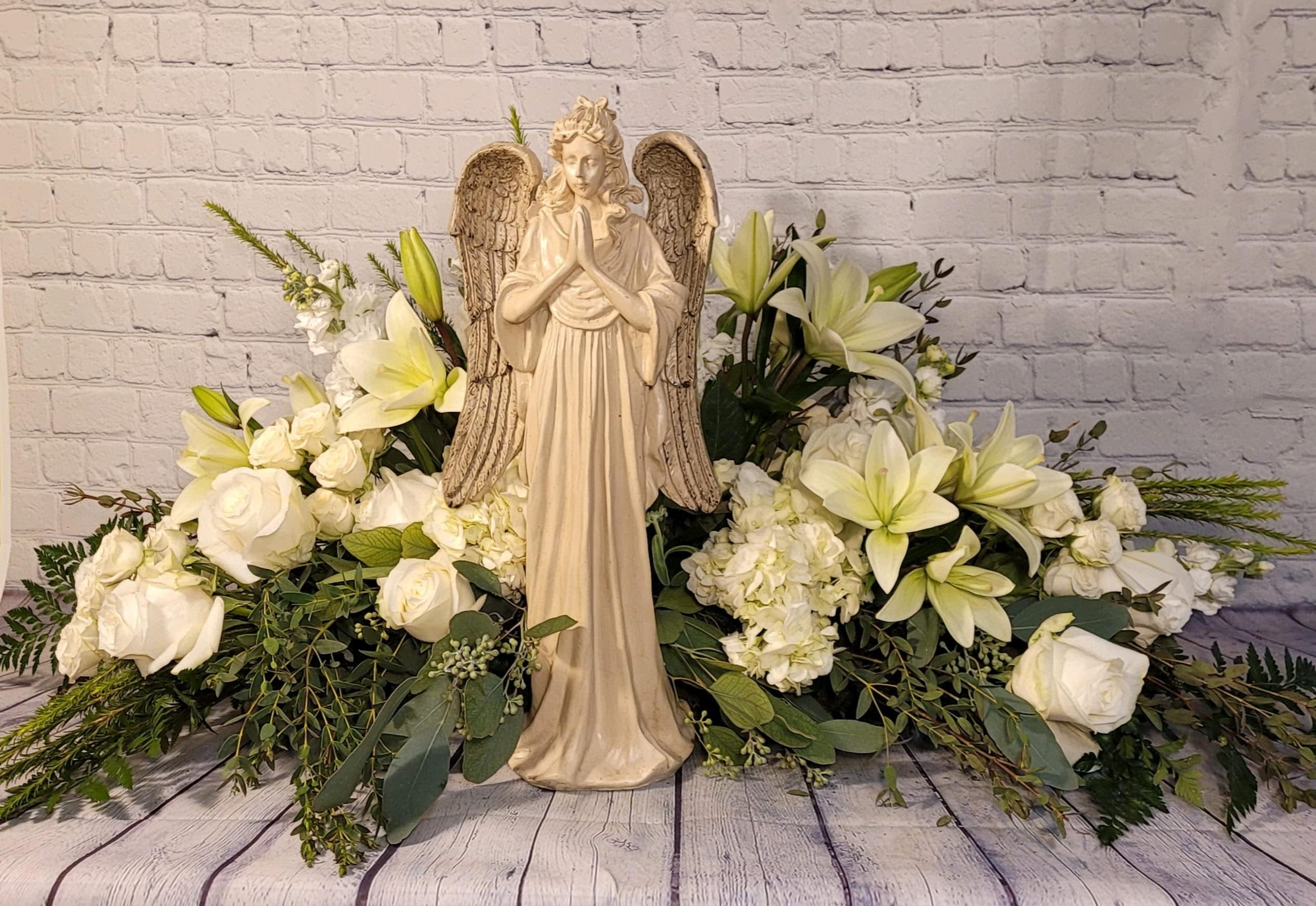 Philia - Philia, the Highest Form of Love. This grand arrangement is centered around a large resin angel figurine and accented with a variety of all white flowers.  Flowers shown; hydrangea, lilies, roses, spray roses, &amp; stock.