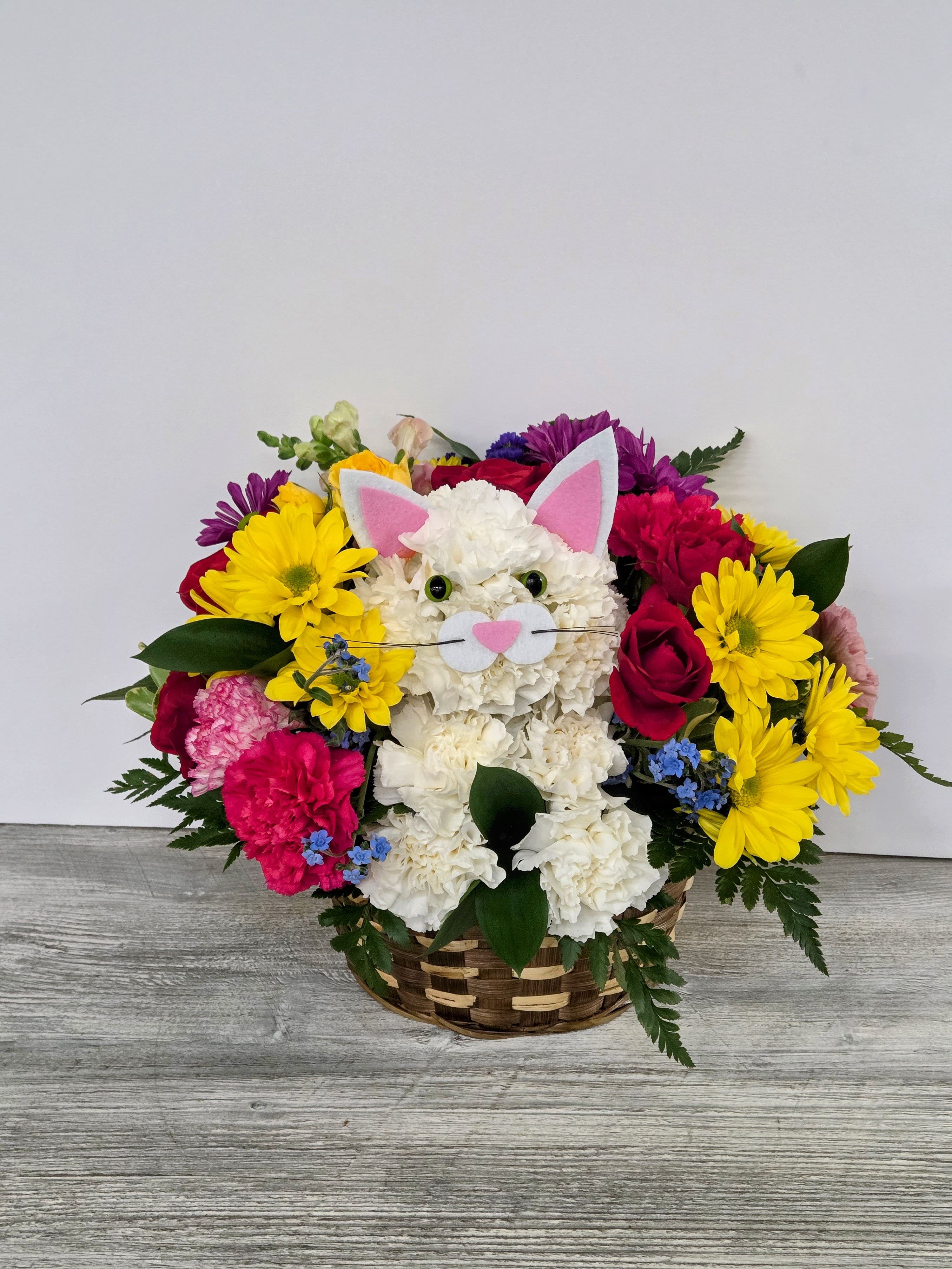Basket arrangement with a white carnation cat face surrounded by yellow daisies, red roses, and bright mixed flowers