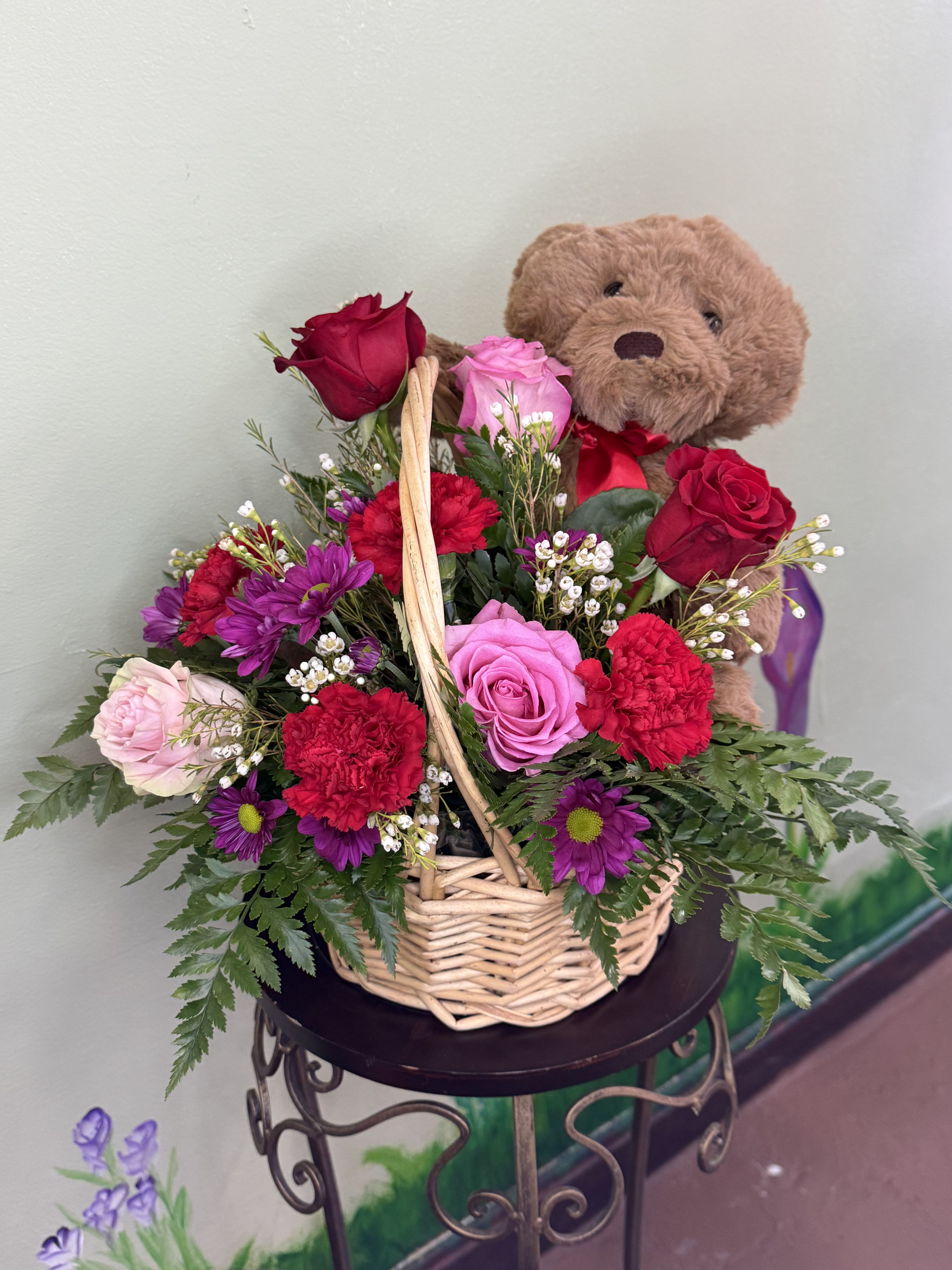 Basket arrangement of red and pink roses, red carnations, purple daisies, and a plush teddy bear.
