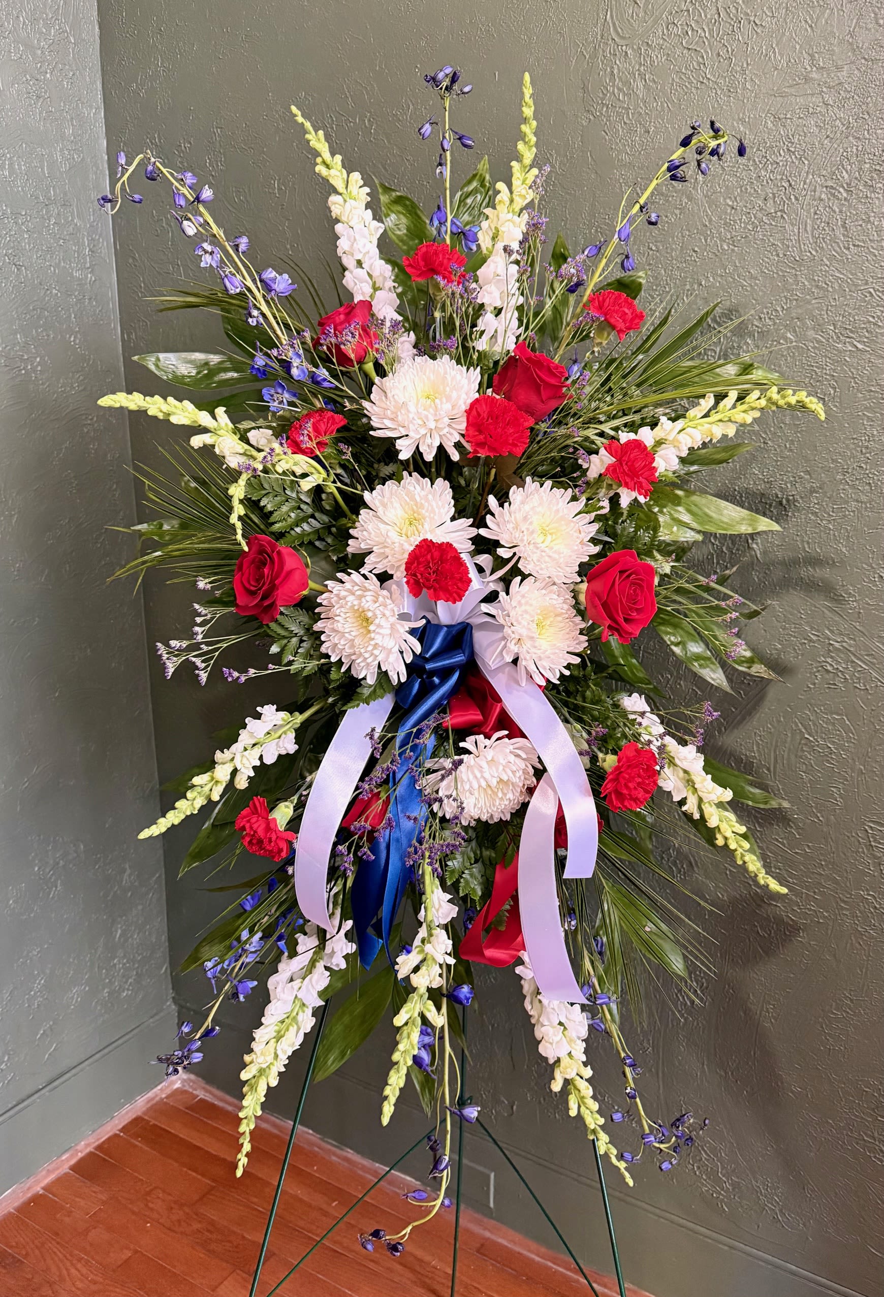 Tall standing spray of red roses, white mums, and blue accents with patriotic ribbons on an easel.