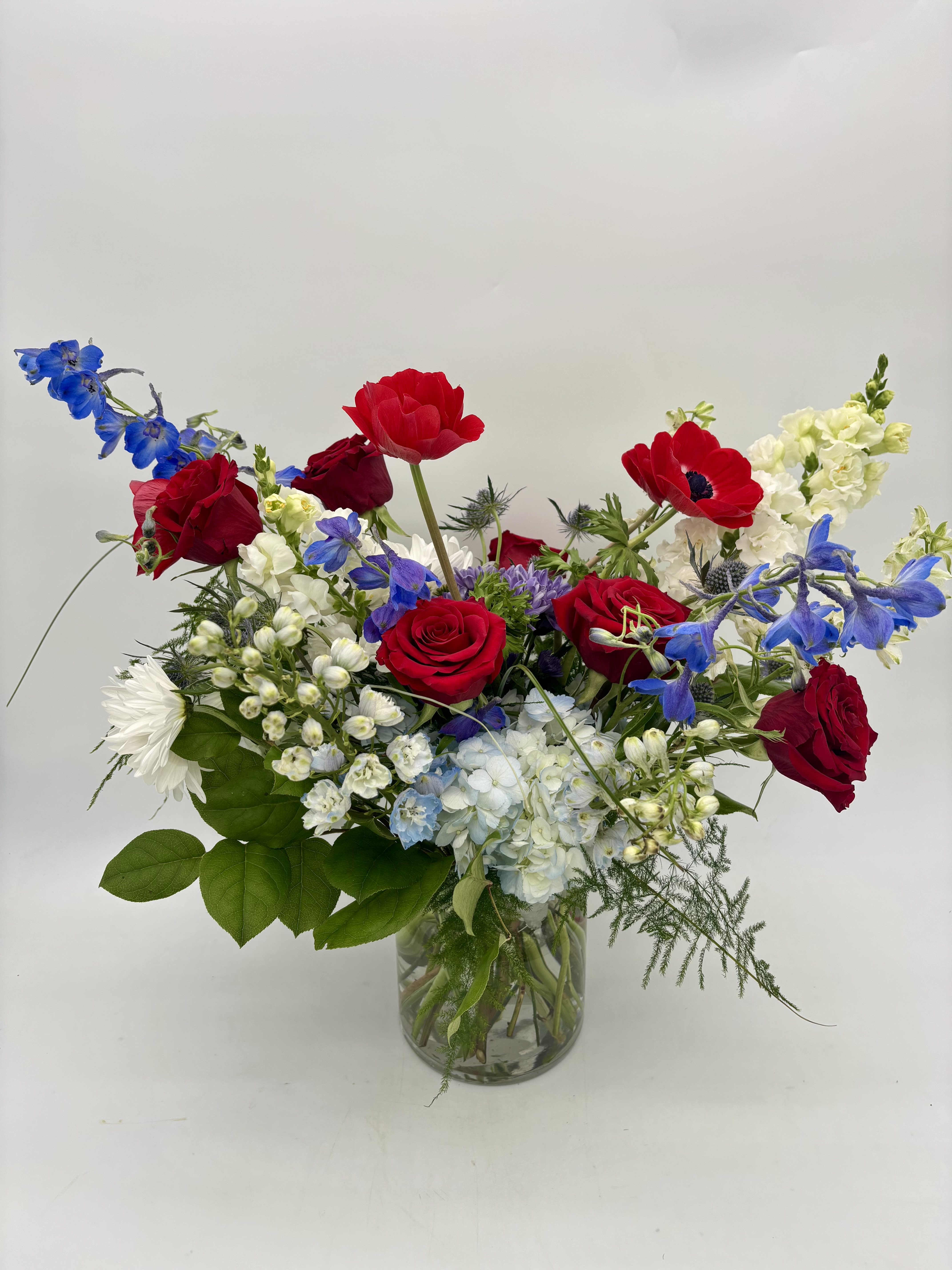 America's Sweetheart - Happy 250 America! A bold celebration of color, America's Sweetheart features red roses and anemones paired with striking blue and is softened by crisp white blooms. Designed with lush greenery in a modern cinched vase, this arrangement brings an elevated patriotic feel with dramatic height and garden-inspired movement.  Perfect for summer celebrations, honoring loved ones, or making a statement with classic Americana colors.