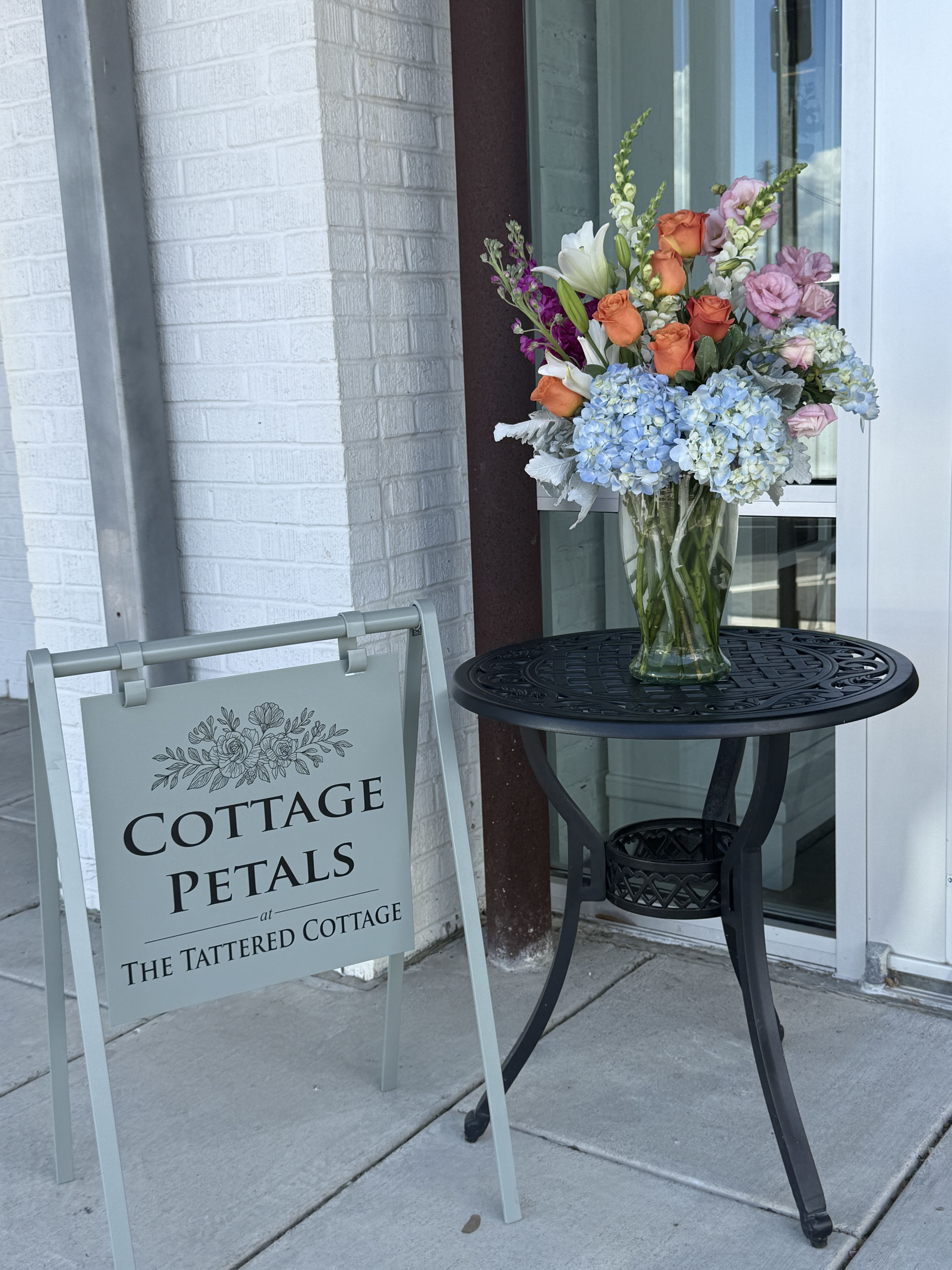 Garden of Gentle Memories - Soft, elegant, and full of grace, the Garden of Gentle Memories arrangement features delicate blue hydrangeas, peach roses, blush blooms, white lilies, and vibrant seasonal accents thoughtfully arranged in a clear glass vase. Designed to bring comfort and peace during difficult times, this arrangement offers a beautiful expression of love, remembrance, and heartfelt sympathy.  Its soft pastel palette and natural garden-inspired style create a warm and comforting tribute suitable for funeral services, memorial gatherings, or sending support to loved ones.  Flower varieties and accent blooms may vary slightly based on seasonal availability, but the overall style, fullness, and soft color palette will always be maintained.
