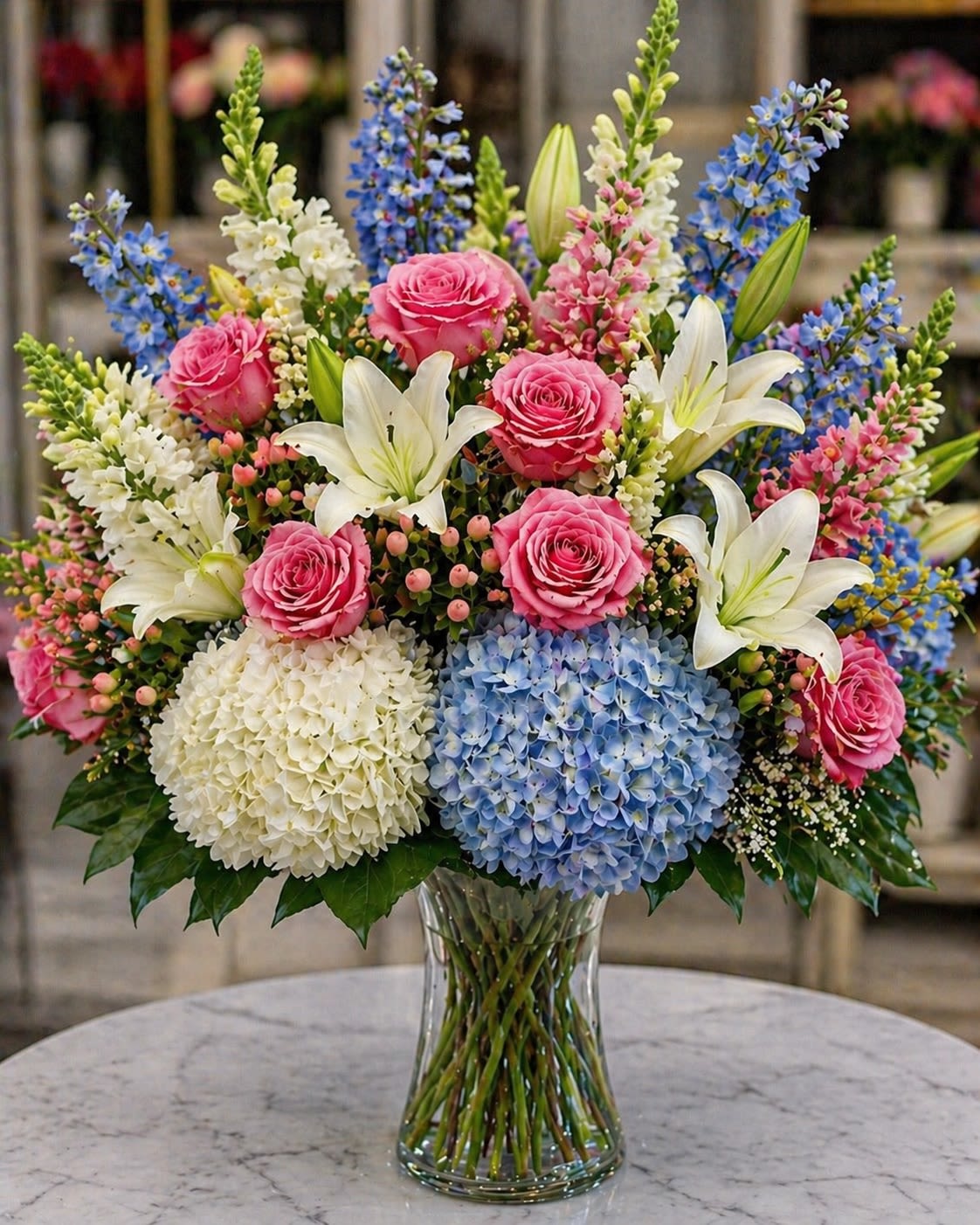 Azure Romance - A lush garden-style floral arrangement featuring soft pink roses, elegant white lilies, blue and white hydrangeas, delicate snapdragons, blooming delphinium, pink wax flowers, and seasonal greenery arranged in a clear glass vase. Azure Romance blends soft pastel romance with airy blue tones, creating a bouquet that feels fresh, graceful, and luxuriously timeless. The delicate balance of pink, white, and powder blue florals gives this design an elegant European garden aesthetic with a dreamy spring feel.