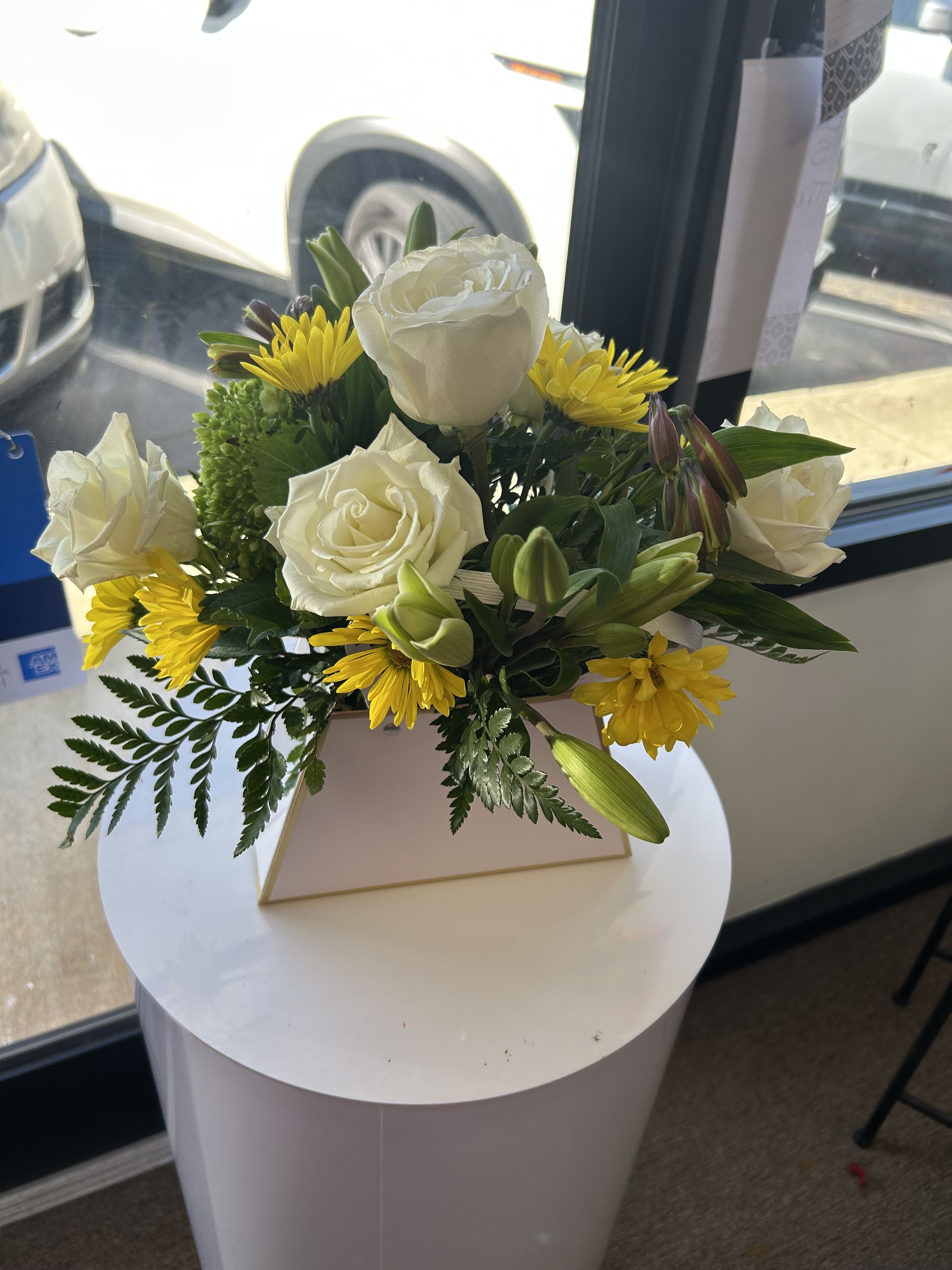 Glam Bag  - Soft white roses meet sunny yellow daisies and poised lily buds in a compact, rounded flower arrangement made for brightening someone's day. This flower arrangement for everyday gifting and cheerful occasions pairs white, yellow, and green tones to create a clean, uplifting look that feels both elegant and optimistic - a lovely choice for birthdays, thank-yous, or "just because" moments.  Arranged tightly in a purse like vase, the bouquet features open white roses, daisy-like yellow blooms, and several unopened lily buds surrounded by deep green foliage that adds contrast and structure. The box keeps the design modern and easy to display on a desk or tabletop; the unopened lilies offer evolving interest as they open over a few days. It's the kind of gift that makes the recipient feel noticed and genuinely cheered - fresh, low-fuss, and ready to brighten any room.