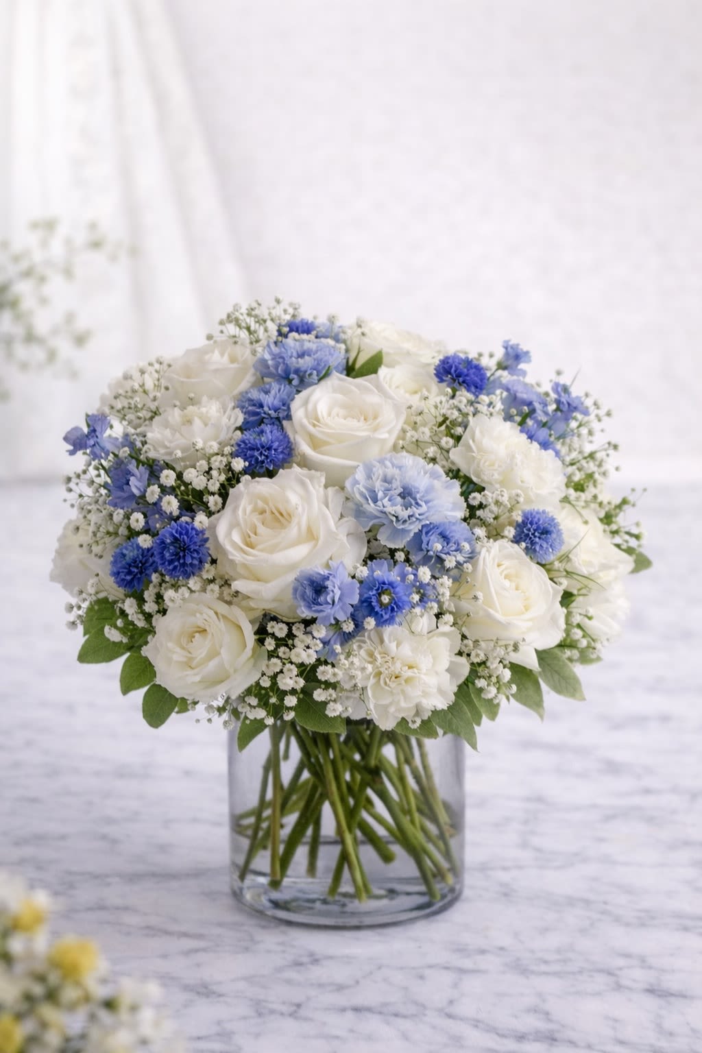 Blue Serenity Bloom - A graceful blend of crisp white roses, soft blue carnations, delicate baby’s breath, and seasonal accent blooms arranged in a clear glass cylinder vase. This design features a calming palette of white and soft blue tones, creating a fresh and serene look that’s perfect for sympathy, new baby celebrations, birthdays, or simply sending comfort and love. The clean glass vase enhances the natural beauty of the blooms while keeping the arrangement timeless and elegant. ⸻  Substitution Policy: Flower varieties may vary based on seasonal availability and market conditions. If substitutions are necessary, blooms will be replaced with flowers of equal or greater value while maintaining the same white and blue color palette and overall fullness of the arrangement. ? Container Notice: Vase style may vary slightly depending on availability, but it will always complement the white and blue color theme. The overall look, feel, and color harmony of the arrangement will remain consistent with the design shown.