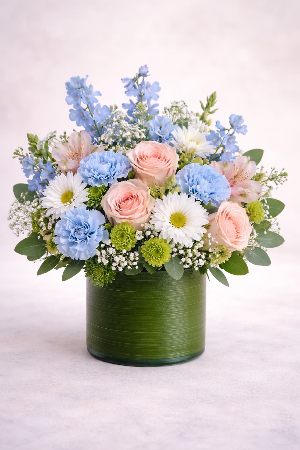 Blue Skies -  Description: A refreshing and elegant arrangement featuring soft blush roses, delicate sky-blue carnations, crisp white daisies, airy baby’s breath, and fresh greenery, beautifully arranged in a modern green cylinder container. This design blends soothing blue tones with romantic blush pink and clean white accents, creating a serene and uplifting look. The balanced mix of soft textures and cool hues makes it perfect for baby showers, birthdays, sympathy, get well wishes, or simply sending a peaceful and thoughtful gesture.  Substitution Policy: Flower varieties may vary based on seasonal availability, but the overall blue, blush, and white color palette will always remain consistent. Any substitutions will be of equal or greater value while preserving the same soft, fresh aesthetic shown. ? Container Notice: Container style may vary slightly depending on availability, but it will always complement the arrangement and maintain the modern, elegant presentation. Our goal is to deliver an arrangement that reflects the same fullness, color harmony, and graceful beauty as pictured.