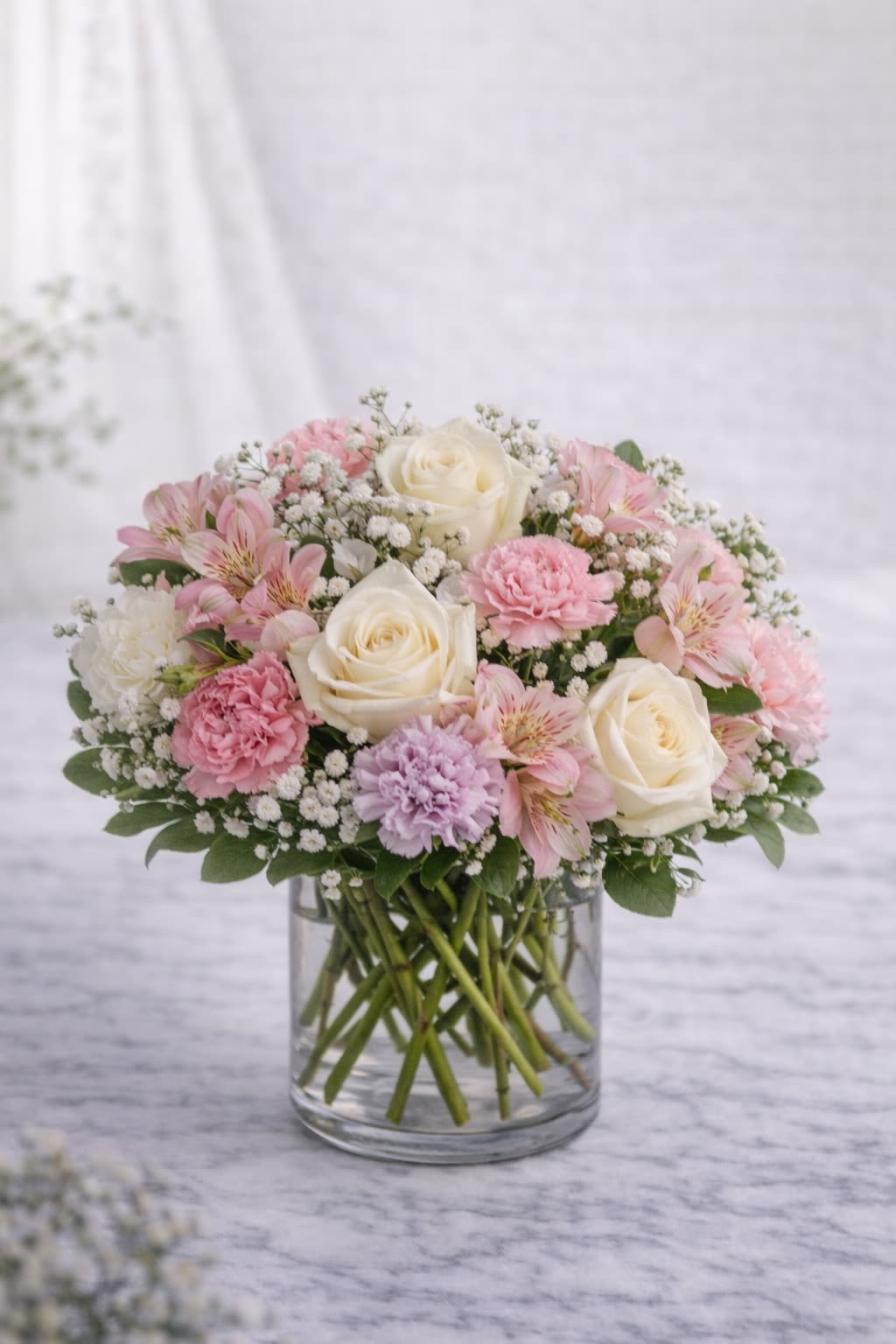 Blush Elegance -  Description: A graceful and romantic arrangement featuring creamy white roses, soft blush carnations, delicate pink alstroemeria, lavender accents, and airy baby’s breath, beautifully arranged in a clear glass vase. Designed in soft pastel tones of ivory, blush pink, and subtle lavender, this arrangement creates a timeless and elegant look. Its rounded, full shape makes it perfect for anniversaries, sympathy, birthdays, baby showers, or simply sending love. Soft. Refined. Effortlessly beautiful. ⸻  Substitution Policy: Due to seasonal availability and market conditions, specific flower varieties may vary. If substitutions are necessary, blooms will be replaced with flowers of equal or greater value while maintaining the same soft pastel color palette (white, blush pink, and light lavender tones). The overall size, fullness, and romantic style of the arrangement will always remain consistent with the design shown. ⸻ ? Container Notice: Vase style may vary slightly based on availability. However, it will always complement the soft, elegant tones of the arrangement. The overall look and color harmony will remain true to the original design.