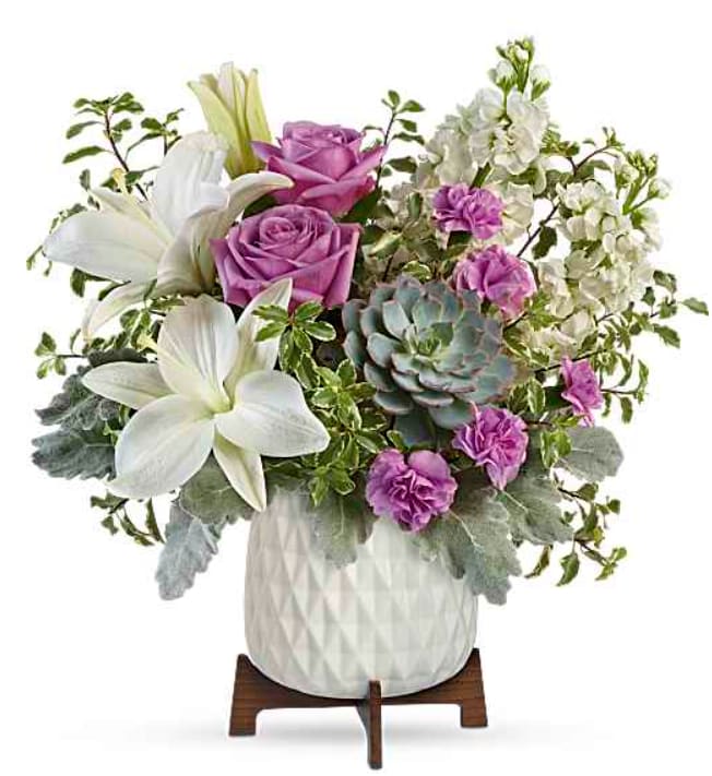 Garden Oasis Bouquet - Soft lavender roses and large white lilies create a calm, contemporary statement in this garden-inspired flower arrangement-perfect as a birthday bouquet, anniversary gift, or thoughtful everyday surprise. The mix of purple and white blooms paired with a striking blue-green succulent gives the person you're thinking of a fresh, modern look that feels both elegant and relaxed.  This arrangement is layered and full in a rounded white ceramic vase set on a wooden stand, with airy white blossoms and clustered purple carnations balancing one another in a slightly asymmetrical composition. Soft gray-green foliage and trailing branches add texture and volume without overpowering the flowers; small pink and white filler blooms bring delicate contrast. The succulent provides a modern texture counterpoint to the soft petals, making the piece decorative enough to read as a centerpiece for home or office.  Send this to someone who deserves to feel seen-whether marking a birthday, saying thank you, offering sympathy, or celebrating an anniversary. Designed for easy display, the bouquet arrives arranged in the ceramic vase on its wooden stand; to keep it looking fresh, change the water every few days and trim stems as needed.