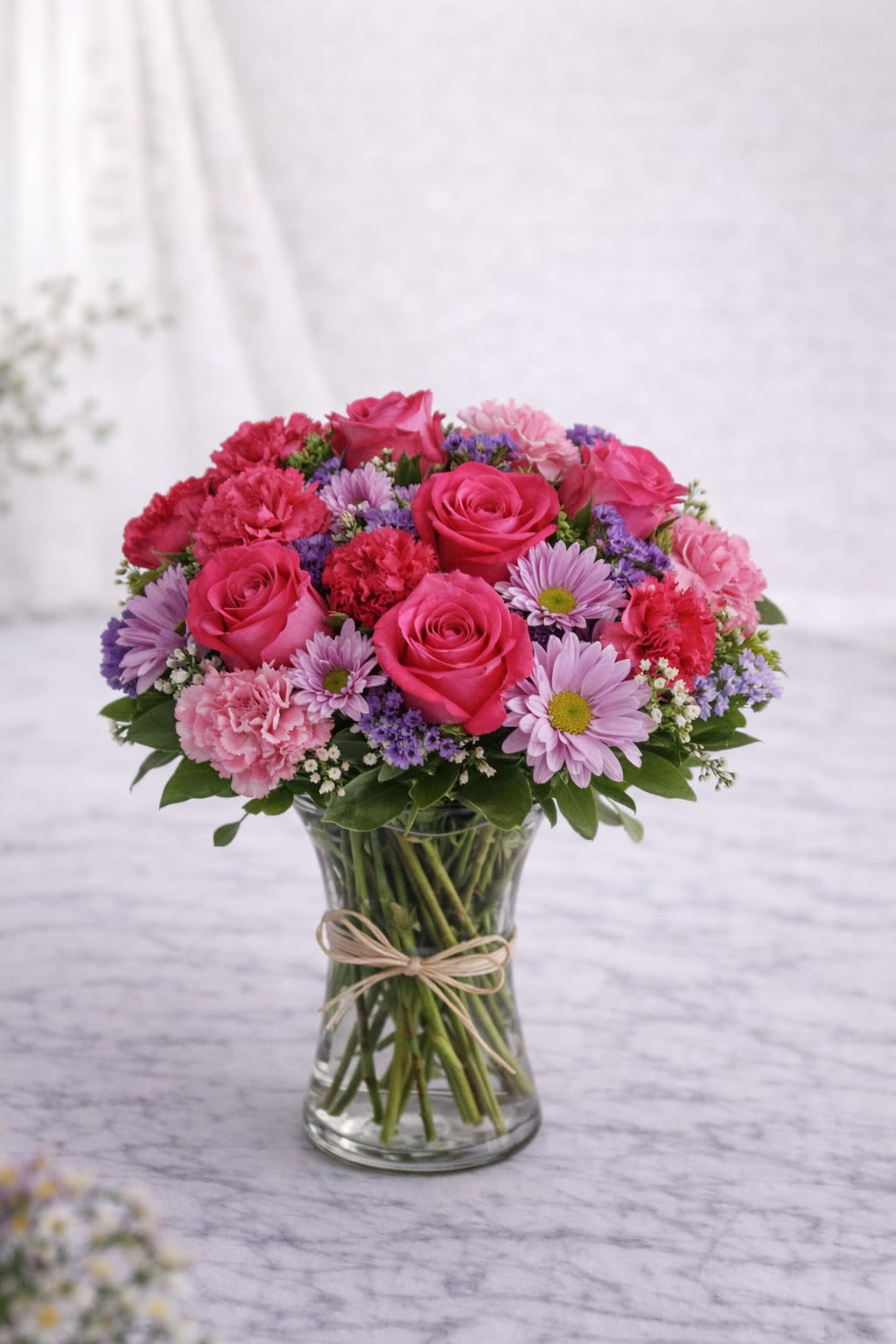 Blushing Romance - Description: A lush and charming blend of hot pink roses, soft blush carnations, lavender daisies, and delicate filler flowers, beautifully arranged in a classic clear glass vase tied with a natural ribbon accent. This design combines rich pink tones with soft lavender and fresh greenery, creating a romantic and joyful look. The rounded, full shape makes it perfect for anniversaries, birthdays, love celebrations, or simply sending a heartfelt surprise. Elegant, feminine, and full of warmth. ⸻  Substitution Policy: Due to seasonal availability and market conditions, specific flower varieties may vary. If substitutions are necessary, blooms will be replaced with flowers of equal or greater value while maintaining the same pink and lavender color palette. The overall size, fullness, and romantic style of the arrangement will always remain consistent with the design shown.