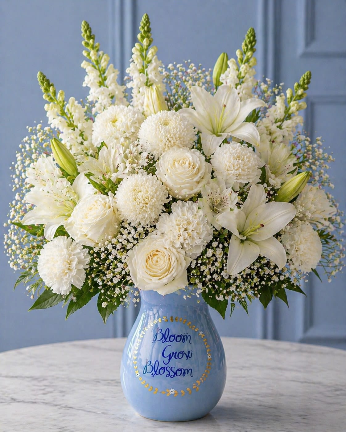 Celestia Blanc - Elegant, airy, and timeless, Celestia Blanc is a luxurious all-white floral composition designed to embody grace, purity, and serenity. Featuring pristine white roses, oriental lilies, chrysanthemums, snapdragons, and delicate baby’s breath arranged in a charming keepsake ceramic vase, this design radiates softness and sophistication. The calming monochromatic palette creates a heavenly aesthetic while layered textures bring depth, fullness, and couture floral styling.