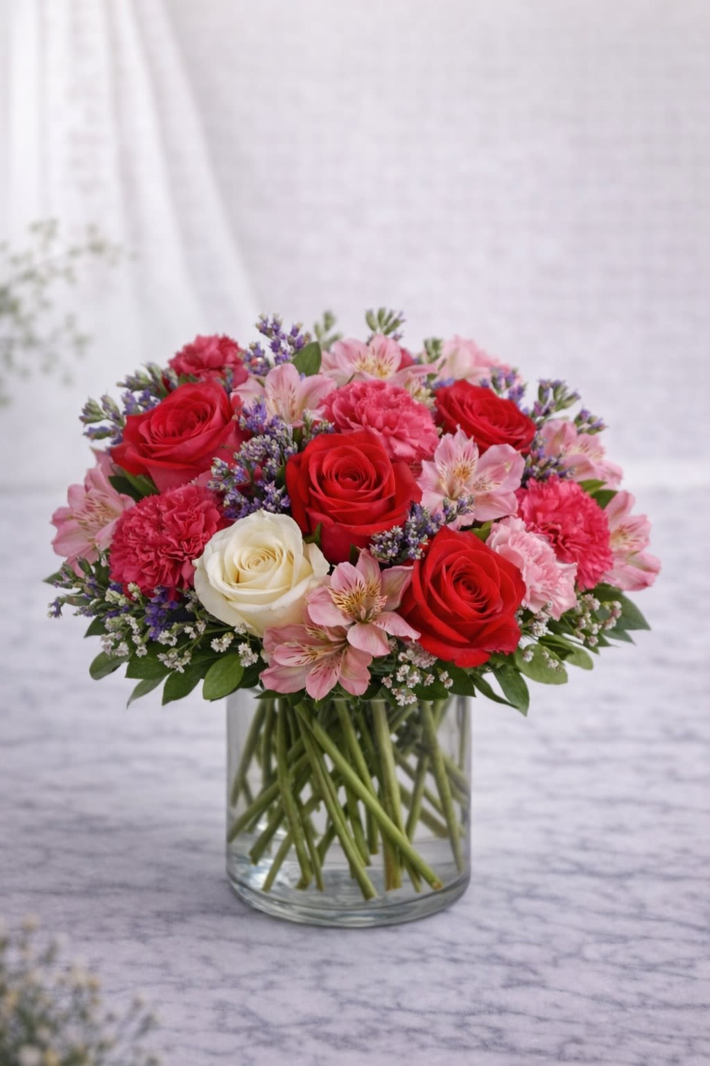 Crimson Romance -  Description: A passionate and elegant arrangement featuring vibrant red roses, soft blush pink blooms, touches of creamy white roses, delicate alstroemeria, and subtle lavender accents, all beautifully designed in a clear glass vase. The rich red tones blend harmoniously with soft pinks and whites, creating a classic romantic color palette that feels both timeless and luxurious. Designed in a full, rounded shape, this arrangement is perfect for anniversaries, romantic gestures, Valentine’s Day, or expressing deep love and admiration. Bold. Romantic. Unforgettable. ⸻  Substitution Policy: Due to seasonal availability and market conditions, specific flower varieties may vary. If substitutions are necessary, flowers will be replaced with blooms of equal or greater value while maintaining the same romantic red, blush pink, and soft white color palette. The overall shape, fullness, and color balance of the arrangement will always remain consistent with the design shown. ⸻ ? Container Notice: Vase style may vary slightly based on availability. However, it will always complement the romantic tones of the arrangement. The overall look and color harmony will remain true to the original design.