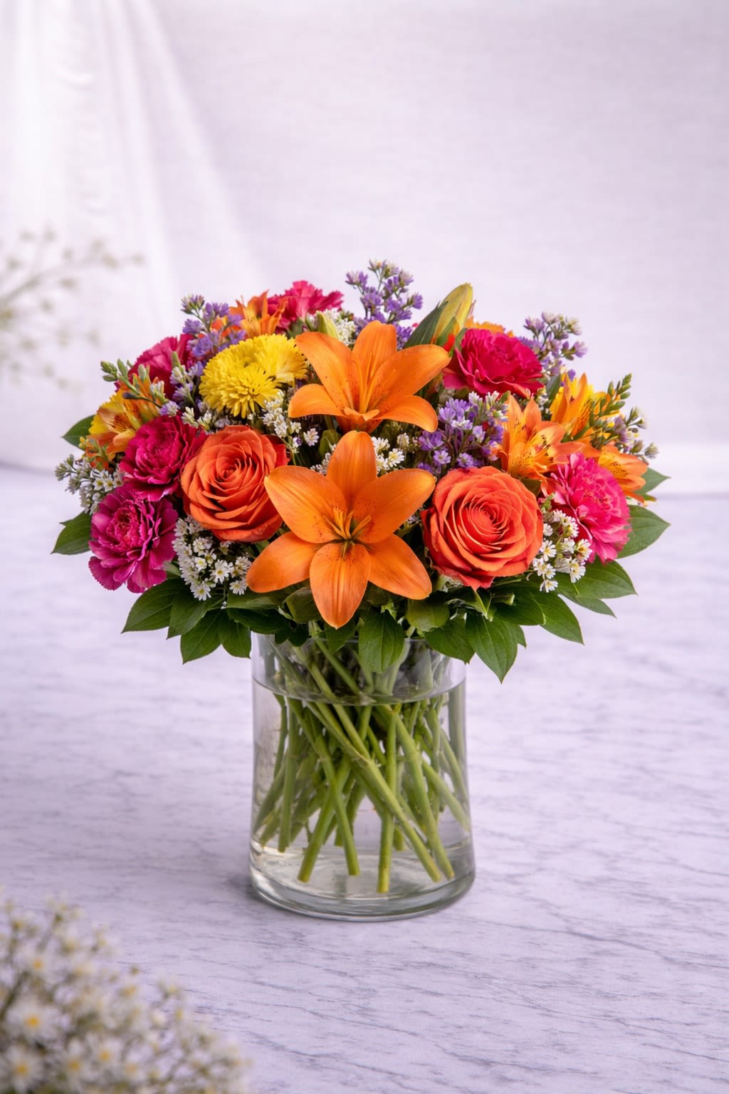 Golden Ember Garden - A bold and radiant arrangement inspired by the warmth of a glowing sunset. Golden lilies take center stage, surrounded by rich orange roses, vibrant pink carnations, and seasonal accent blooms layered with fresh greenery for a full, lush look. The design blends warm tones of amber, coral, and sunshine yellow to create a piece that feels joyful, energetic, and unforgettable. Perfect for birthdays, celebrations, thank-you gifts, or simply brightening someone’s day. Substitution Policy: Flower varieties may vary based on seasonal availability, but the overall warm, sunset-inspired color palette will always remain consistent. Any substitutions will be carefully selected to maintain the same fullness, style, and balanced design. The container will always coordinate beautifully with the arrangement’s colors and overall look.*