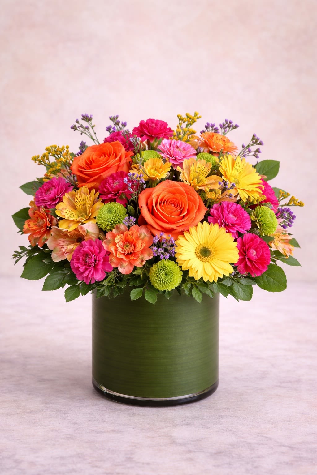 Golden Fiesta - A vibrant and joyful arrangement bursting with warm oranges, bright yellows, lively pinks, and fresh green accents. Designed in a modern green cylinder vase, this piece features radiant roses, cheerful gerbera daisies, playful carnations, alstroemeria, and seasonal fillers for added texture and fullness. The bold color combination creates an energetic, happy feel—perfect for birthdays, congratulations, or simply making someone smile. Substitution Policy: Flower varieties may vary based on seasonal availability, but the bright sunset-inspired color palette of orange, yellow, pink, and green will always remain consistent. Containers may vary slightly in shade or finish while always complementing the overall vibrant design.*
