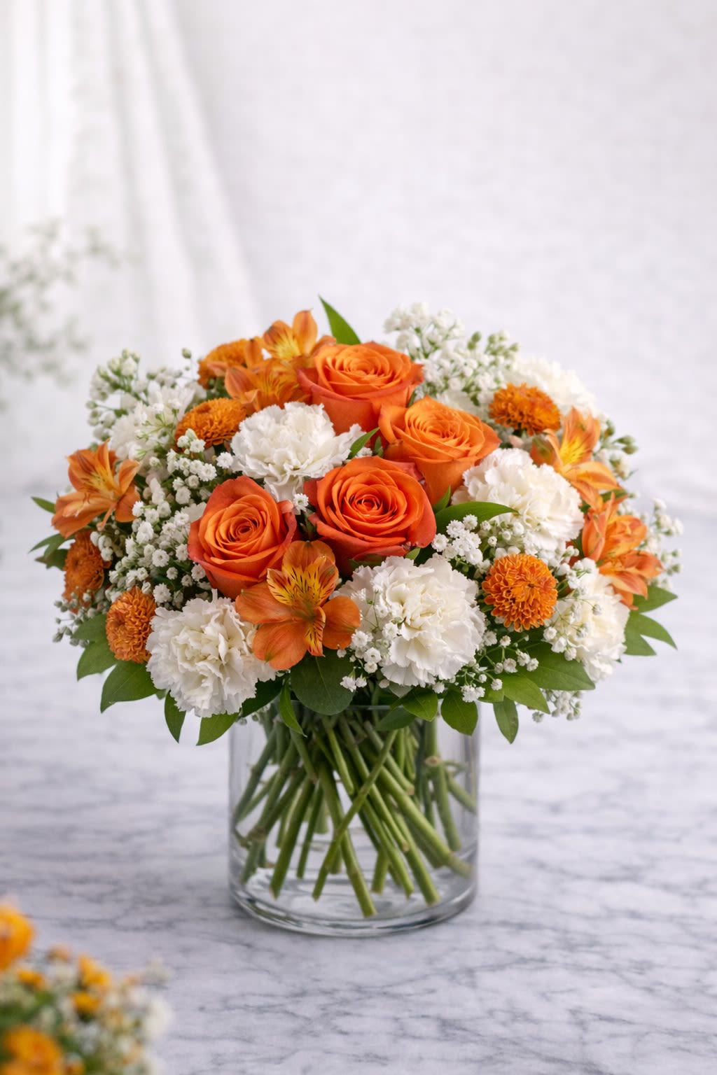 Golden Glow Bouquet Orange - A radiant blend of vibrant orange roses, soft white carnations, delicate alstroemeria, and seasonal accent blooms, beautifully arranged in a clear glass cylinder vase. This design combines warm sunset oranges with fresh whites and touches of green, creating a cheerful and uplifting arrangement that instantly brightens any space. Perfect for birthdays, congratulations, thank you gifts, or sending a little sunshine to someone special. The clear glass vase showcases the natural beauty of the stems, giving this bouquet a clean, modern, and timeless look. ⸻  Substitution Policy: Flower varieties may vary based on seasonal availability and market conditions. If substitutions are necessary, blooms will be replaced with flowers of equal or greater value while maintaining the same vibrant orange and crisp white color palette. The overall style, fullness, and warm sunny feel of the arrangement will always remain consistent with the design shown. ⸻ ? Container Notice: Vase style may vary slightly depending on availability, but it will always complement the orange and white tones of the arrangement. The overall look and color harmony will remain the same.