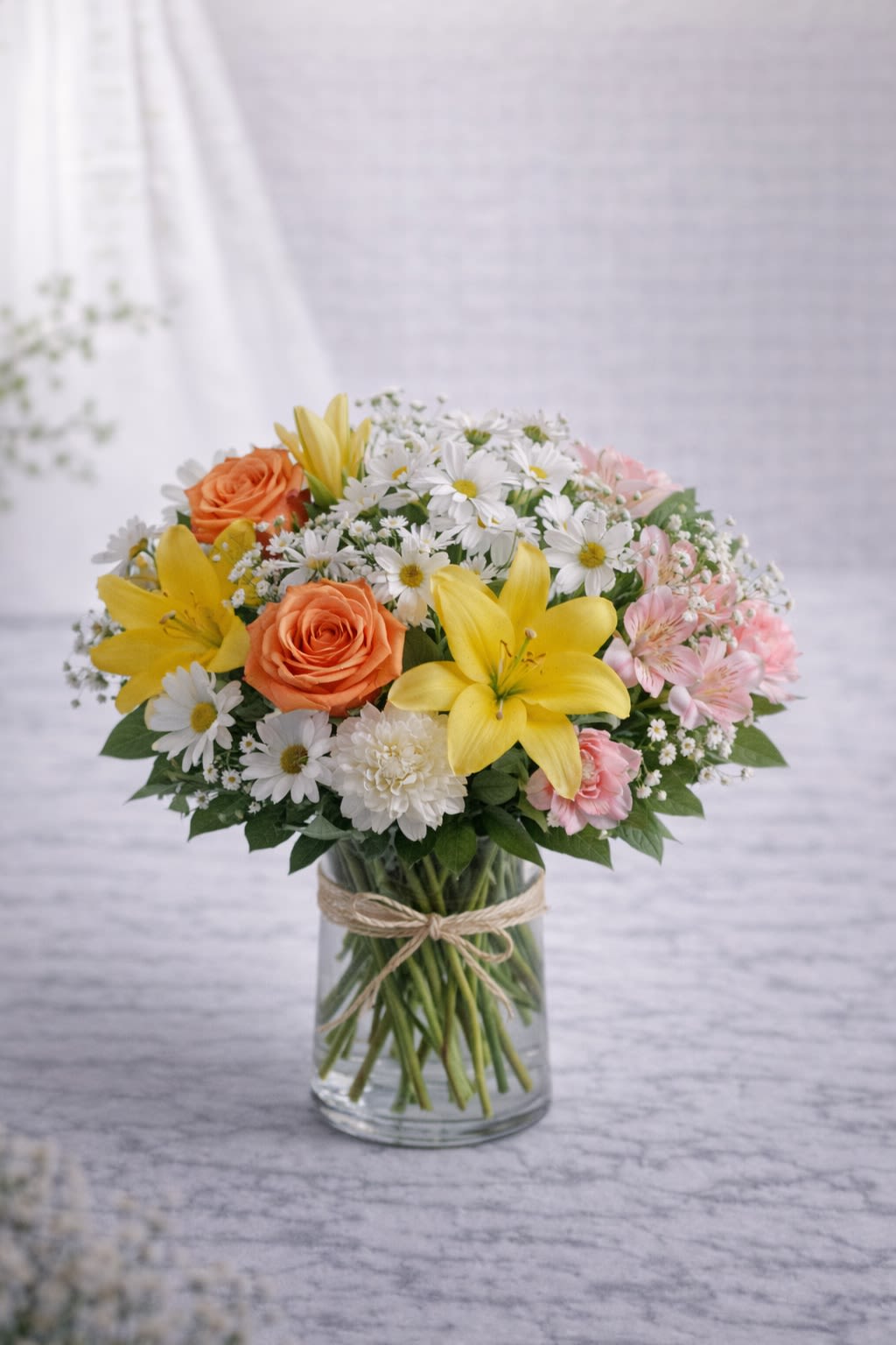 Golden Morning Bliss - Golden Morning Bliss is a radiant and refreshing arrangement inspired by the warmth of sunshine and soft spring air. Featuring bright yellow lilies, peach roses, classic white daisies, delicate pink alstroemeria, and lush greenery, this design feels cheerful, light, and effortlessly elegant. The combination of golden yellows, soft peach, crisp white, and blush pink creates a balanced and uplifting palette—perfect for birthdays, get well wishes, thank-you gifts, or simply bringing joy into someone’s day. Arranged in a clear glass vase with a natural twine accent, it offers a fresh and timeless presentation. Please note: Flower varieties may vary based on seasonal availability. If substitutions are necessary, we will always maintain the same color palette and overall look of the arrangement. Containers and accents will complement the design shown and remain consistent in style and tone. 
