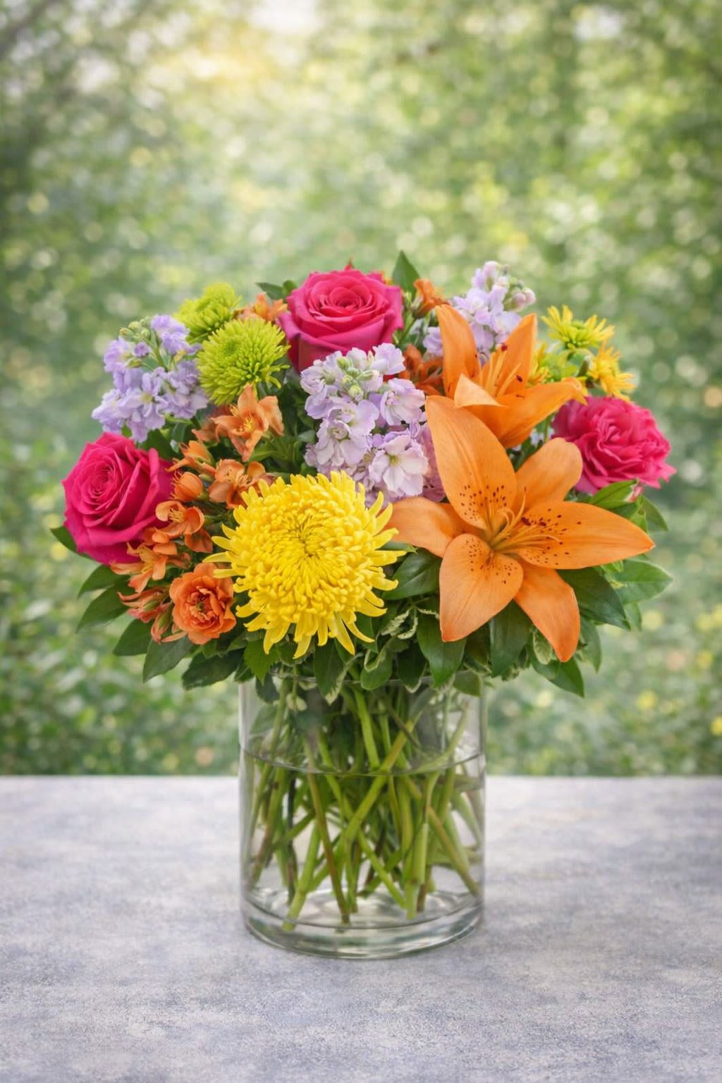 Golden Radiance - A vibrant and joyful arrangement bursting with bold color and fresh energy. Radiant orange lilies and golden chrysanthemums take center stage, surrounded by hot pink roses, soft lavender blooms, and lush greenery. Designed in a clear glass vase, this bouquet feels like a warm summer afternoon — bright, uplifting, and full of life. Perfect for celebrations, thank-you gifts, or simply bringing a pop of happiness into any space, this arrangement makes a statement with its rich color contrast and lively texture. Substitutions: Flower varieties may vary based on seasonal availability, but the bright, multicolor palette and overall fullness will always be maintained. Any substitutions will be carefully selected to preserve the vibrant, cheerful style of the design.* 