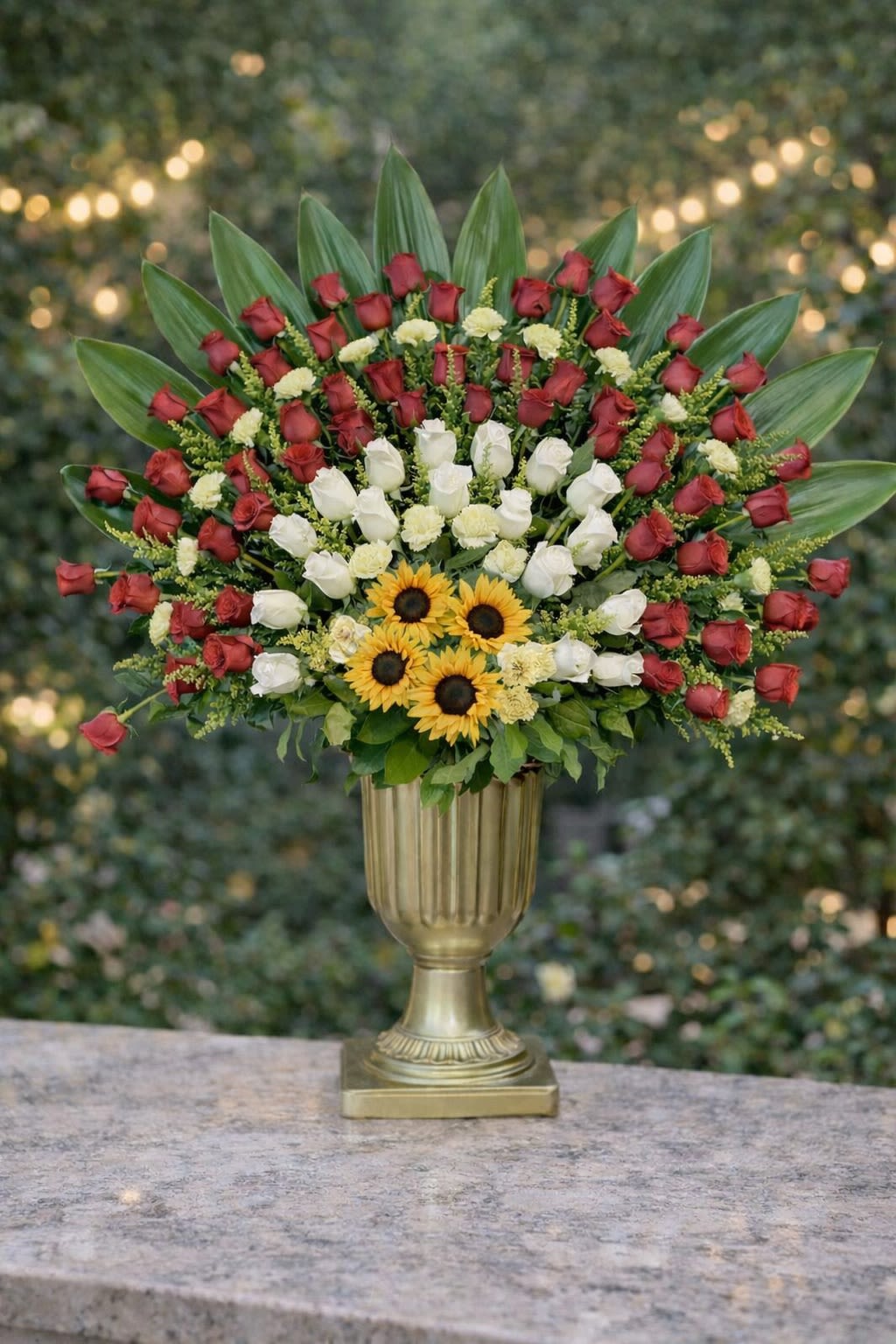 Imperial Rose Symphony - A breathtaking and grand-scale arrangement featuring premium red and white roses, vibrant sunflowers, lush greenery, and dramatic tropical foliage arranged in an elegant gold pedestal vase. Designed in a wide, fan-style silhouette, this piece creates a powerful and luxurious visual impact. Layers of rich crimson roses blend with soft ivory blooms and bold sunflowers, framed by structured greenery for depth and dimension. This arrangement is ideal for milestone celebrations, grand romantic gestures, memorial tributes, weddings, corporate events, or any occasion that calls for an unforgettable floral statement. Seasonal &amp; Freshness Guarantee We carefully source the freshest blooms available to ensure quality, vibrancy, and longevity. Because flowers are seasonal and market availability may vary, certain accent blooms or greenery may occasionally be substituted. If substitutions are necessary, we will: • Maintain the same red, white, and yellow color palette • Preserve the same grand, fan-style structure • Keep the same overall size and premium value • Use flowers and greenery of equal or greater quality. Your arrangement will always reflect the same bold and elegant aesthetic shown — never a reduced or simplified version. Each piece is designed to maintain structure, balance, and dramatic impact.