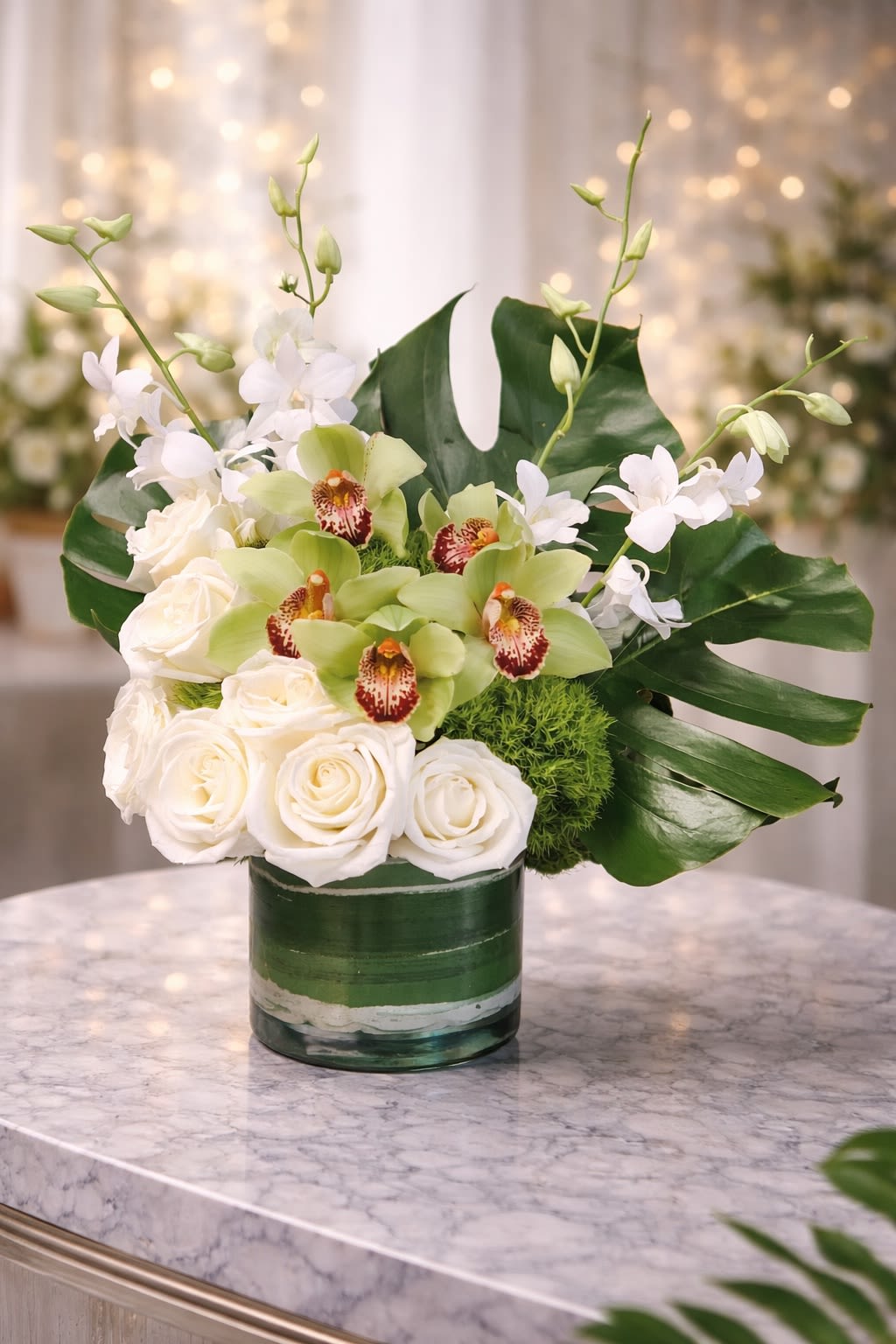 Ivory Orchid Elegance - A refined and luxurious arrangement featuring premium green cymbidium orchids, crisp white roses, delicate dendrobium orchids, and modern tropical foliage. Designed in a sleek glass cylinder with layered greenery, this piece blends soft ivory tones with rich emerald accents for a timeless and sophisticated look. Perfect for anniversaries, sympathy gestures, birthdays, corporate gifting, or elegant home décor. • Premium green cymbidium orchids • Fresh white roses • White dendrobium orchids • Textural green accents • Modern glass vase included This arrangement delivers a clean, upscale aesthetic with balanced height and fullness. ⸻  Seasonal &amp; Freshness Guarantee We carefully select the freshest blooms available to ensure exceptional quality and longevity. Due to seasonal availability, certain flowers or greens may occasionally be substituted. If a substitution is needed, we will: • Maintain the same elegant white and green color palette • Preserve the modern, luxury design style • Ensure the arrangement remains full and balanced • Use flowers of equal or greater value and quality Your order will always reflect the same sophisticated look and feel shown — never a different style or theme. Each arrangement is handcrafted with attention to detail to ensure beauty and consistency.