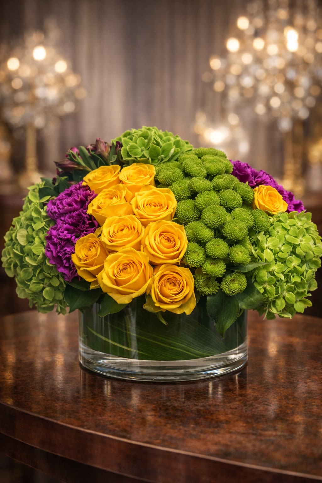 Modern Harvest Glow - A bold and contemporary arrangement featuring vibrant golden roses, lush green hydrangea, textured green button blooms, and rich purple accent flowers arranged in a sleek glass cylinder vase. Designed in a structured, rounded style, this piece blends warm golden tones with fresh greens and deep jewel accents for a modern and eye-catching presentation. The layered textures create depth and fullness while maintaining a clean, upscale aesthetic. Perfect for birthdays, congratulations, thank-you gifts, corporate occasions, or brightening any space with vibrant color. • Premium golden roses • Fresh green hydrangea • Textured green button blooms • Purple accent florals • Modern glass cylinder vase included Handcrafted for fullness, symmetry, and luxury presentation. ⸻  Seasonal &amp; Freshness Guarantee We carefully select the freshest blooms available to ensure vibrant color and lasting beauty. Because flowers are seasonal and availability may vary, certain accent blooms or specific shades may occasionally be substituted. If substitutions are necessary, we will: • Maintain the same yellow, green, and purple color palette • Preserve the same full, modern rounded design • Keep the same overall size and premium value • Use flowers of equal or greater quality Your arrangement will always reflect the same bold and contemporary aesthetic shown — never a different theme or reduced design. Each piece is handcrafted with attention to density, balance, and elegance.