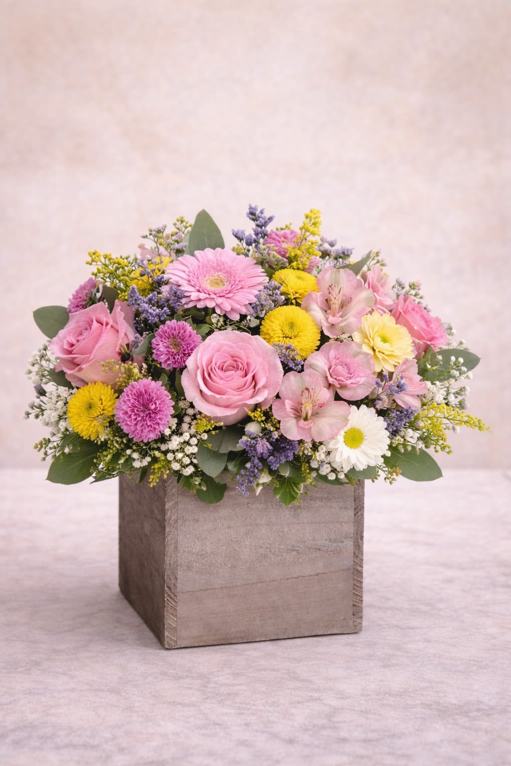 Pastel Radiance - A cheerful yet elegant arrangement designed in a rustic wooden cube, featuring soft pink roses, delicate gerbera daisies, yellow button mums, alstroemeria, and textured seasonal fillers. The blend of blush pinks, sunny yellows, and gentle lavender accents creates a bright and uplifting look while maintaining a soft, romantic feel. Perfect for birthdays, thank-you gifts, or simply bringing light into someone’s day. Substitution Policy: Flower varieties may vary based on seasonal availability, but the pastel pink and yellow color palette and overall balanced, full design will always remain the same. The wooden container may vary slightly in tone or style, while always complementing the arrangement’s soft and cheerful aesthetic.*