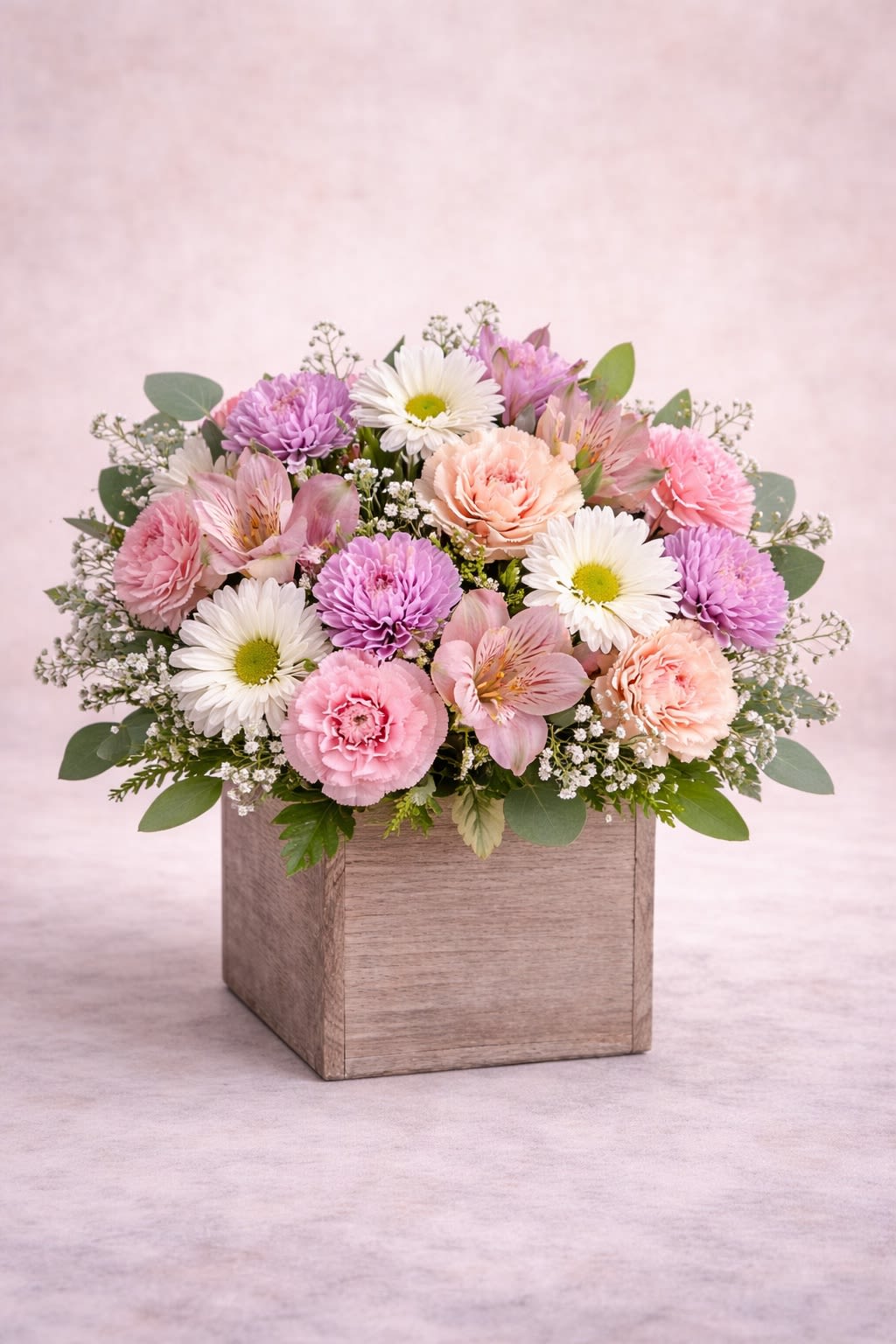 Pastel Serenity Box -  Description: A soft and elegant arrangement featuring delicate pink carnations, creamy blush blooms, lavender cushion mums, white daisies, and airy accent flowers, beautifully designed in a modern wooden box container. This design blends gentle pastel pinks, soft lavenders, and crisp whites to create a calming and sophisticated look. The combination of textures and subtle color contrasts makes it perfect for birthdays, get well wishes, sympathy gestures, baby showers, or simply to brighten someone’s day.  Substitution Policy: Flower varieties may vary depending on seasonal availability, but the overall pastel color palette and balanced, garden-inspired design will always remain consistent. Any substitutions will be of equal or greater value while preserving the soft and elegant aesthetic shown. ? Container Notice: Wood box style may vary slightly based on availability, but it will always complement the arrangement and maintain the same modern, natural presentation. Our goal is to deliver an arrangement that reflects the same fullness, harmony, and delicate beauty as pictured.