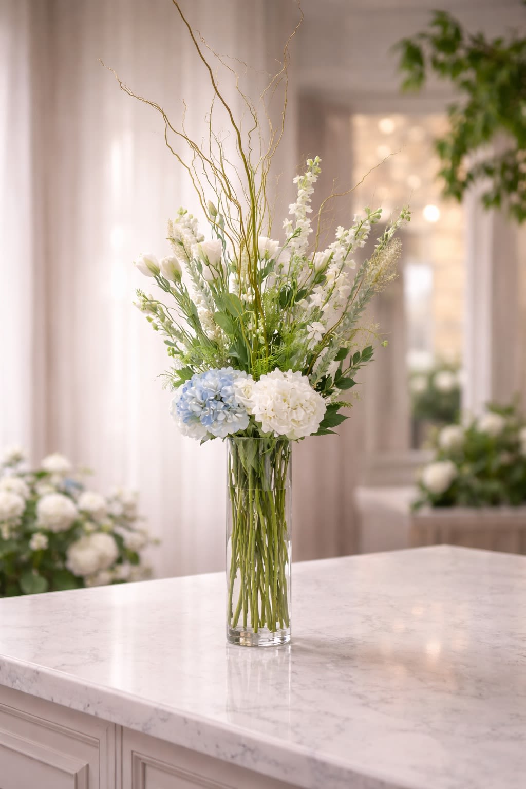 Serene Sky Tower - A graceful and elevated arrangement featuring soft white blooms, delicate seasonal florals, and lush hydrangeas accented with natural curly willow branches for height and movement. Designed in a tall, clear glass vase, this piece blends crisp whites, soft blue tones, and fresh greenery for an airy and sophisticated presentation. The vertical design creates a light, flowing silhouette that adds elegance and dimension to any space. Perfect for sympathy, memorial services, weddings, baby showers, corporate settings, or refined home décor. • Premium white hydrangeas • Soft blue hydrangea accents • Delicate seasonal white blooms • Natural curly willow branches • Tall clear glass vase included • Elegant vertical design Handcrafted to create height, balance, and a refined statement. ⸻  Seasonal &amp; Freshness Guarantee We carefully select the freshest seasonal blooms to ensure longevity and beauty. Because flowers and branch elements are seasonal, certain accent blooms or branch varieties may occasionally be substituted. If substitutions are necessary, we will: • Maintain the same white and soft blue color palette • Preserve the same tall, airy, vertical design style • Keep the same overall size, fullness, and premium value • Use flowers and accents of equal or greater quality. Your arrangement will always reflect the same elegant and refined aesthetic shown — never a different theme or reduced style. Each piece is thoughtfully designed to maintain height, flow, and balance.