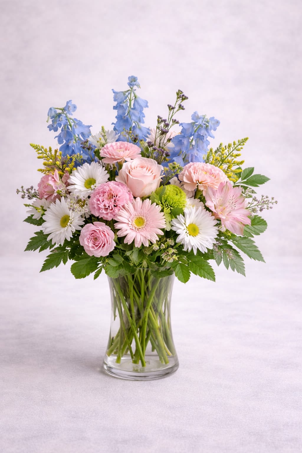 Skylight Serenity -  Description: A soft and airy arrangement featuring blush pink roses, delicate pink gerberas and carnations, crisp white daisies, fresh greenery, and tall light blue delphinium for a graceful touch of height. Designed in a clear glass vase, this bouquet feels fresh, bright, and effortlessly elegant. The gentle blend of pastel pinks, clean whites, and calming sky blues creates a peaceful and uplifting look — perfect for birthdays, new babies, sympathy, get well wishes, or simply brightening someone’s day. The balanced textures and soft color harmony give it a timeless, refreshing charm.  Substitution Policy: Flower varieties may vary based on seasonal availability, but the overall pastel pink, white, and soft blue color palette will always remain consistent. Any substitutions will be of equal or greater value while preserving the same light, airy design style. ? Container Notice: Vase style may vary slightly depending on availability, but it will always complement the arrangement and maintain the elegant, fresh aesthetic shown. Our goal is to deliver a bouquet that reflects the same softness, fullness, and serene beauty as pictured.