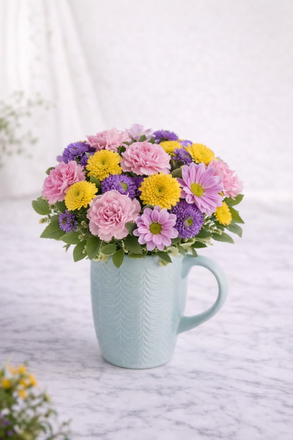 Sunshine Garden Mug - A joyful mix of soft pink carnations, lavender and yellow daisy poms, and seasonal accent blooms, beautifully arranged in a reusable pastel ceramic mug. This design features a playful blend of pink, purple, and sunny yellow tones, creating a fresh, uplifting arrangement that’s perfect for birthdays, get well wishes, thank you gifts, or just because. The keepsake mug adds a charming touch and can be reused long after the flowers are enjoyed. ⸻  Substitution Policy: Flower varieties may vary based on seasonal availability and market conditions. If substitutions are necessary, blooms will be replaced with flowers of equal or greater value while maintaining the same pink, purple, and yellow color palette and overall fullness of the design. ? Container Notice: Mug style and texture may vary slightly depending on availability, but it will always coordinate with the arrangement’s cheerful color theme. The overall look, feel, and color harmony will remain consistent with the design shown.