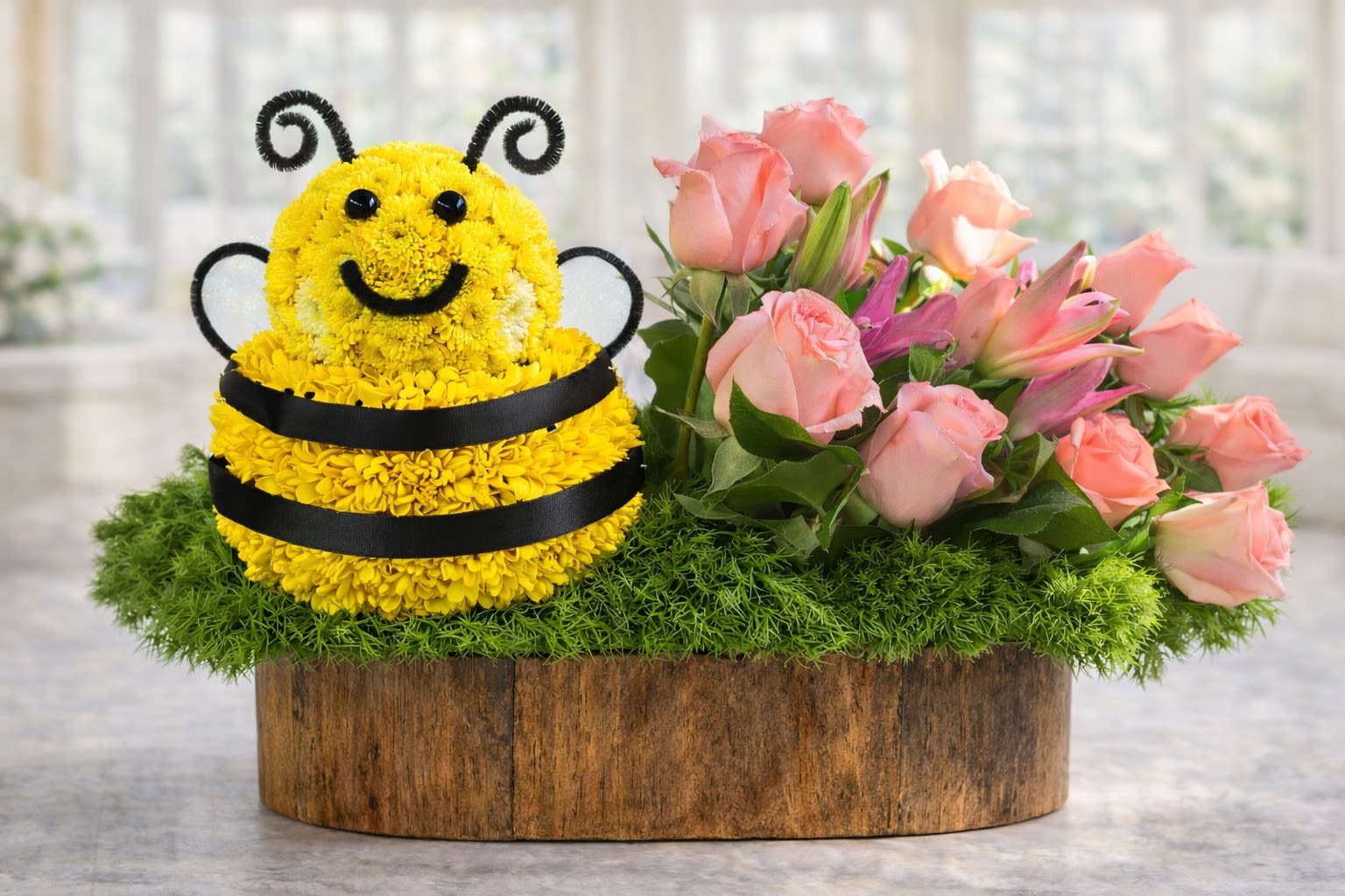 Sweet Bee Garden - A charming and playful floral arrangement featuring an adorable handcrafted bee design created with fresh yellow blooms, paired with soft pink roses, elegant lilies, lush greenery, and moss accents beautifully arranged in a rustic wooden container. This cheerful design is perfect for birthdays, baby showers, spring celebrations, get well wishes, or simply bringing a smile to someone special.  Flower varieties and container style may vary slightly based on seasonal availability and freshness. In the rare event certain flowers or containers are unavailable, substitutions of equal or greater value will be used while maintaining the overall design style and color palette shown.