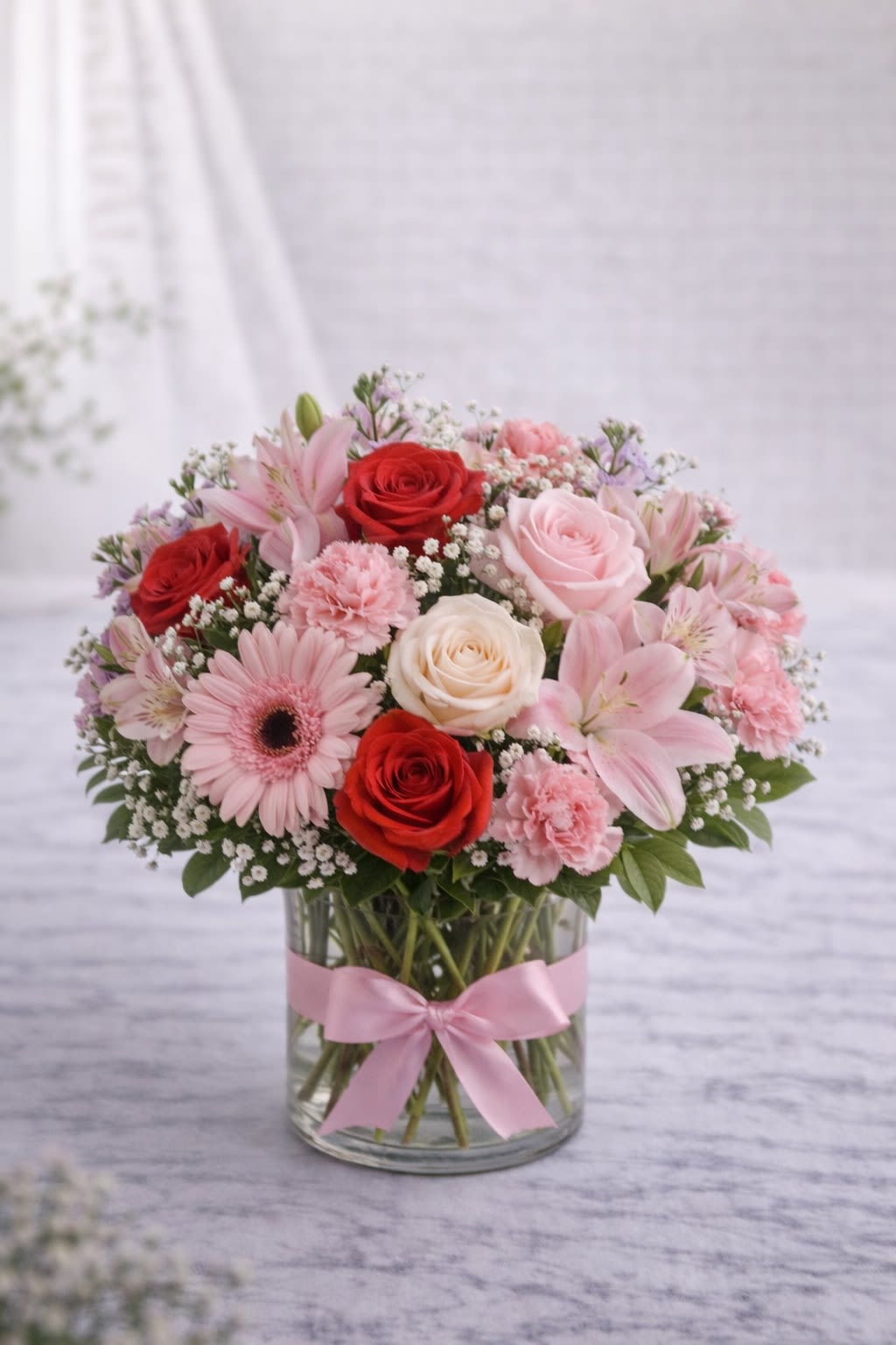 Sweetheart Symphony - Sweetheart Symphony is a charming blend of romantic reds, soft blush pinks, and creamy whites, artfully arranged in a clear glass vase accented with a delicate ribbon. Featuring roses, lilies, gerbera daisies, carnations, and airy filler flowers, this design creates a lush, rounded presentation full of texture and elegance. This arrangement is perfect for anniversaries, romantic occasions, birthdays, or simply to say “I’m thinking of you.” The mix of bold red roses and soft pink tones gives it a timeless, heartfelt feel. Please note: Flower varieties may vary depending on seasonal availability. If substitutions are necessary, we will always maintain the same color palette and overall style while using flowers of equal or greater value. Containers and ribbon styles may vary slightly, but they will always complement the arrangement’s colors. The overall look and color harmony will remain consistent with the design shown. 