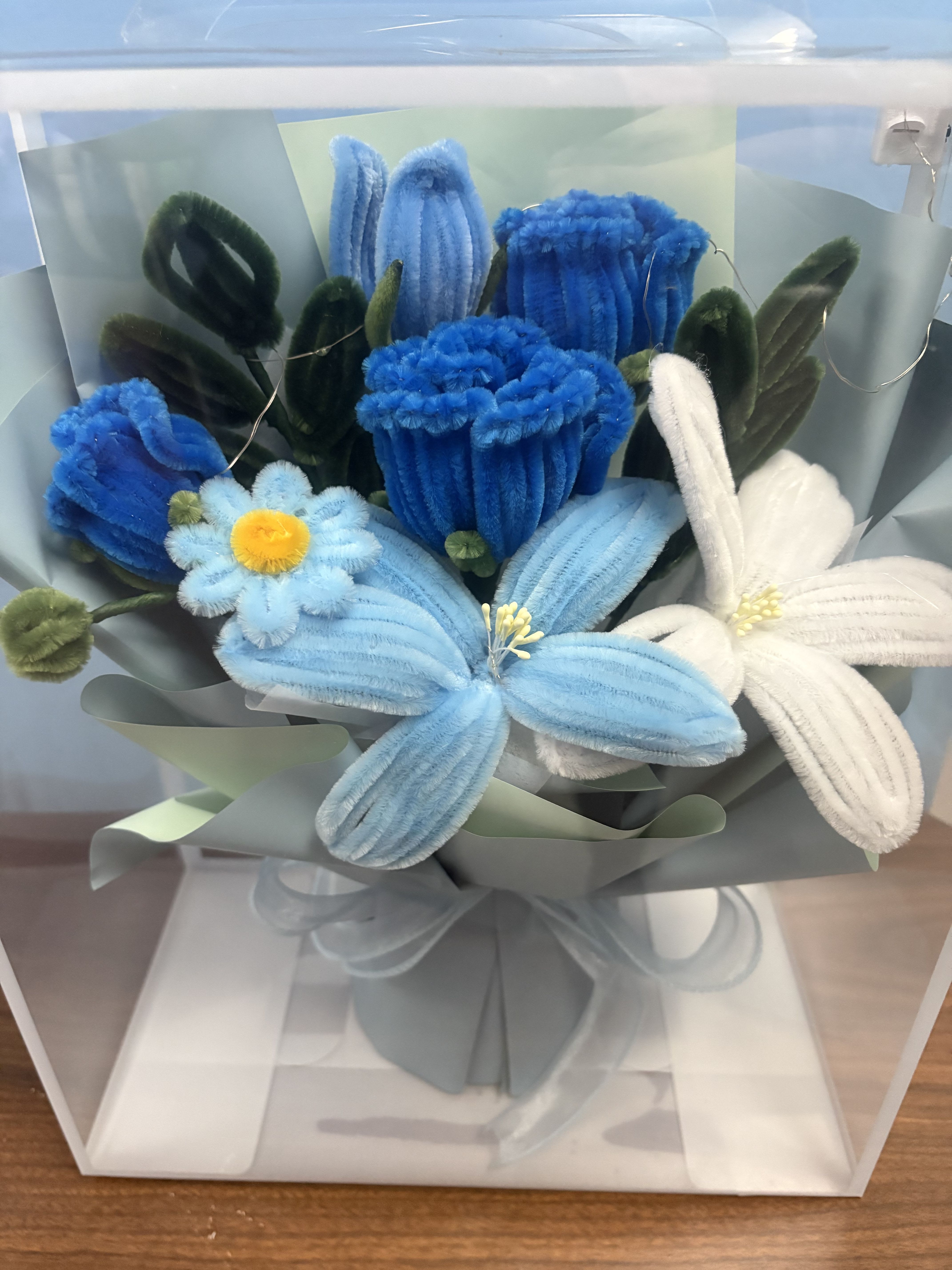  Chenille Bouquet Blue - Soft, playful chenille blooms make this blue bouquet an ideal everyday gift or casual celebration arrangement that feels handcrafted. As a cheerful birthday bouquet or a simple "thinking of you" surprise, the plush pipe-cleaner-style flowers in bright and pale blues with touches of white read as whimsical and fun-she'll feel genuinely seen by a gift that looks made just for her.  The compact arrangement pairs fuzzy blue, pale blue, and white blooms (one with a small yellow center) with dark green fuzzy accents for contrast. It's wrapped in layered pastel or gray paper, tied with a soft ribbon, and offered in options including a clear gift bag or a neat clear display box; a vase option is also available for tabletop display. Lightweight and decorative rather than perishable, it's a keepsake-style bouquet that brightens a desk, shelf, or bedside table without any maintenance.