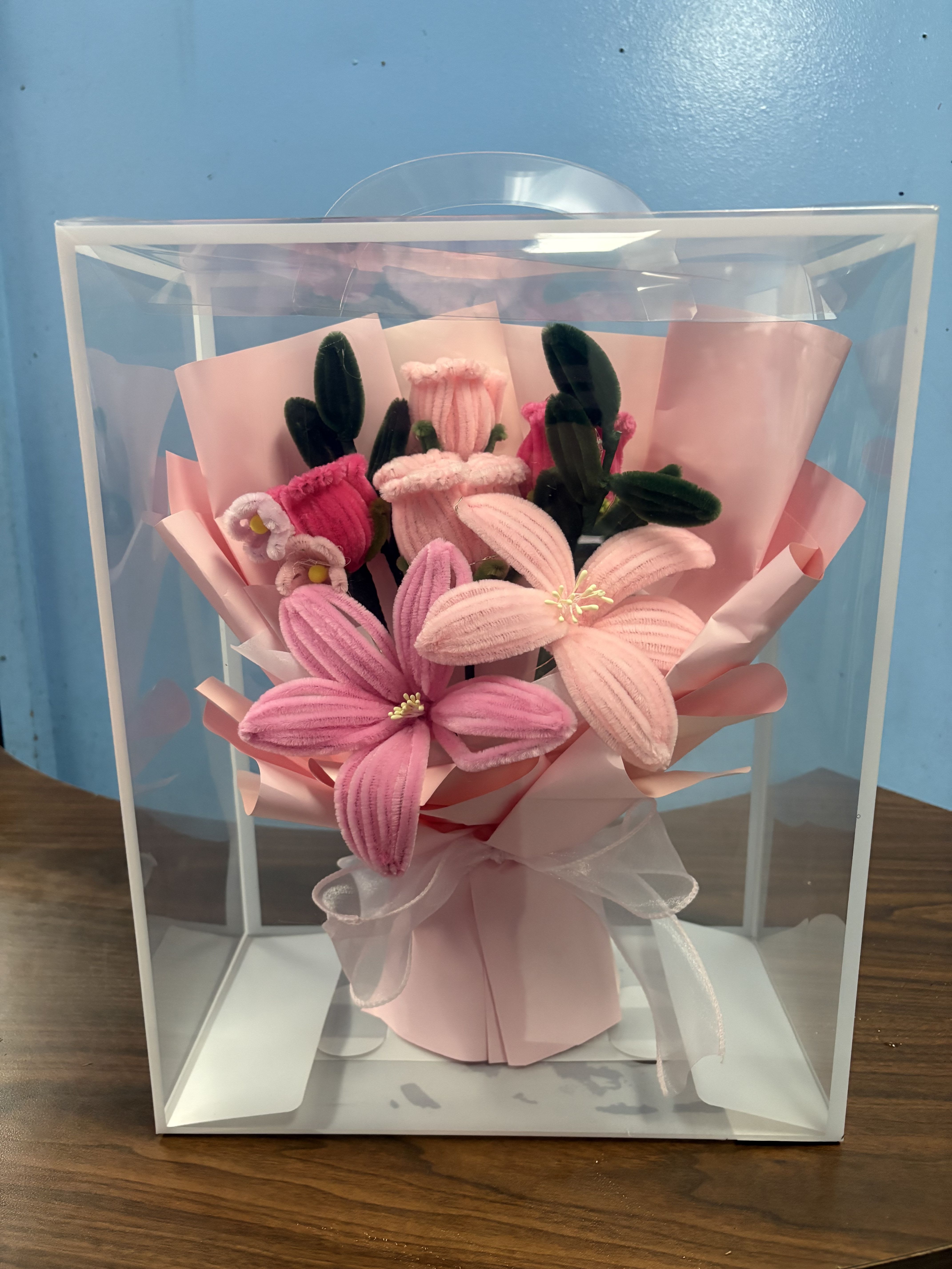  Chenille - Plush ribbed blossoms in soft pink make an immediately cheerful impression - a petite flower arrangement perfect for a birthday bouquet, a "thinking of you" gift, or a small anniversary surprise. The handcrafted, playful texture of the chenille-like petals and the gentle white accents give the recipient a sweet, romantic feeling without the need for fresh-flower care.  This arrangement is made from silk and other artificial materials and is presented in pink wrapping paper inside a clear display box. Dark green bud- and leaf-like accents add contrast to the rounded, hand-tied composition, and a sheer white ribbon is tied around the base for a delicate finishing touch. Compact and decorative, it arrives ready to display in its clear gift box - ideal for sending a lasting token that feels thoughtful and fuss-free.