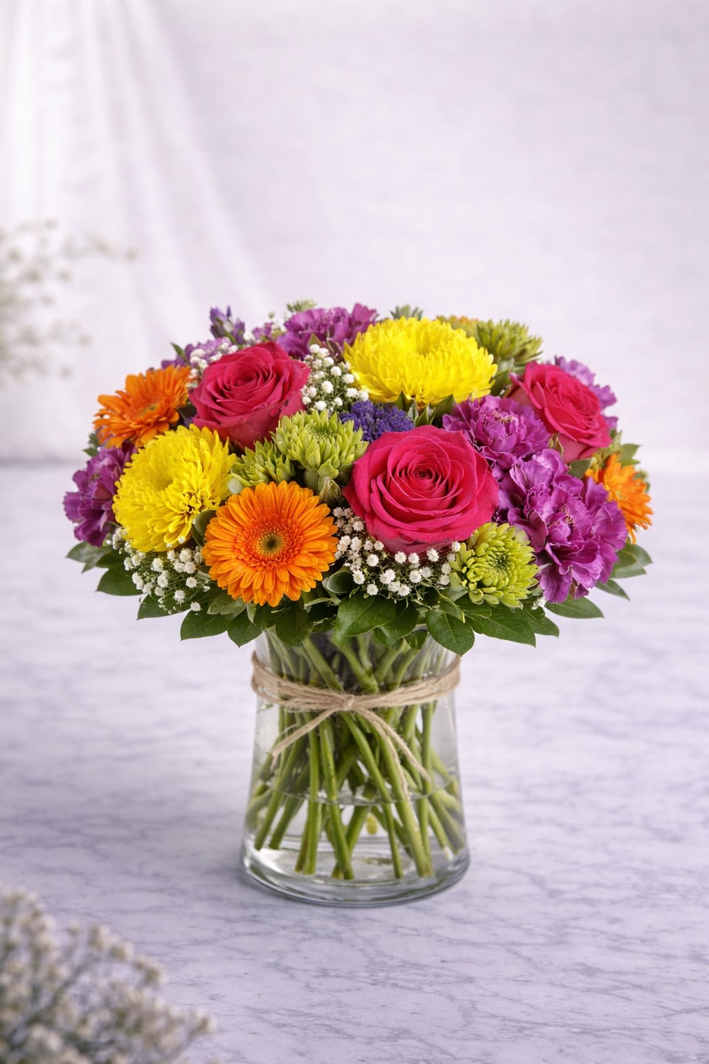 Vibrant Celebration - Vibrant Celebration is a joyful burst of bold color designed to brighten any space instantly. This lively arrangement features hot pink roses, golden yellow chrysanthemums, bright orange gerbera daisies, rich purple carnations, and fresh green accents, beautifully arranged in a clear glass vase wrapped with rustic twine. Full of texture and energy, this design is perfect for birthdays, congratulations, thank-you gifts, or simply to surprise someone with something cheerful and uplifting. The mix of warm and cool tones creates a balanced yet eye-catching display that feels festive and modern. Please note: Flower varieties may vary depending on seasonal availability. If substitutions are necessary, we will maintain the same vibrant color palette and overall style while using flowers of equal or greater value. Container style and decorative accents may vary slightly but will always complement the design shown. 