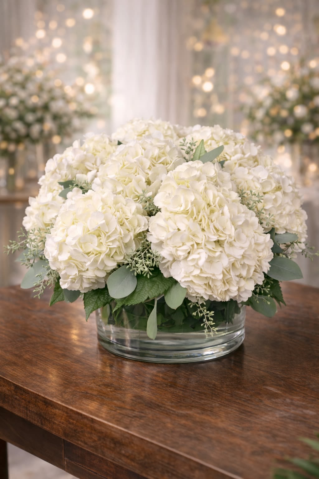 White Clouds - A timeless and elegant arrangement featuring lush premium white hydrangeas accented with soft greenery in a modern clear glass vase. Designed in a full, rounded style, this piece creates a cloud-like effect with soft ivory tones and natural texture. The simplicity of all-white hydrangeas makes it incredibly versatile and sophisticated. Perfect for sympathy, weddings, anniversaries, corporate gifts, housewarmings, or minimalist home décor. • Premium white hydrangeas • Fresh seasonal greenery accents • Modern low glass vase included • Full, rounded luxury design This arrangement is rich, clean, and beautifully balanced. ⸻  Seasonal &amp; Freshness Guarantee We carefully select the freshest hydrangeas available to ensure fullness and longevity. Because hydrangeas are seasonal and natural products, bloom size may vary slightly depending on availability. If needed, we may substitute greenery accents to maintain freshness. If any substitutions are required, we will: • Maintain the same all-white color palette • Preserve the same full, rounded design • Keep the same overall size and premium value • Use blooms of equal or greater quality Your arrangement will always reflect the same elegant, clean aesthetic shown — never a different color theme or reduced style. Each piece is handcrafted to ensure density, balance, and beauty.