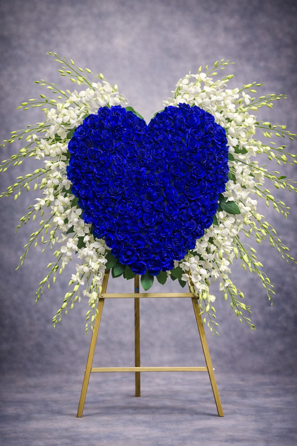 Eternal Blue Heart - A striking heart-shaped standing tribute designed with deep blue roses and accented with cascading white florals and lush greenery. This elegant arrangement symbolizes eternal love, remembrance, and devotion.  Displayed on an easel, this heartfelt design creates a meaningful presence at funeral services, memorials, and celebration-of-life gatherings.  Substitutions may be necessary based on seasonal availability, but the overall heart shape, color intensity, and fullness will always be maintained.  Delivery available to funeral homes, churches, and residences. Please include service time and location details at checkout.