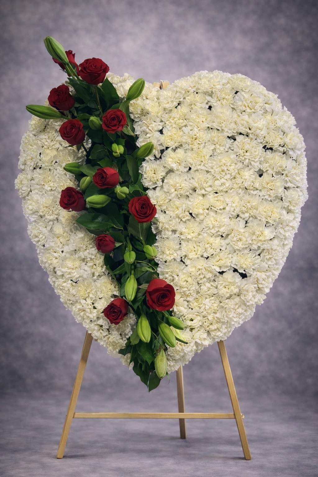 Pure Devotion Heart - A timeless heart-shaped standing tribute designed with soft white carnations and accented with a striking cascade of red roses and fresh greenery. This elegant arrangement symbolizes eternal love, respect, and deep devotion.  Displayed on an easel, this heartfelt design creates a meaningful presence at funeral services, memorials, and celebration-of-life gatherings.  Substitutions may be necessary based on seasonal availability, but the overall heart shape, fullness, and color contrast will always be maintained.  Delivery available to funeral homes, churches, and residences. Please include service time and location details at checkout.