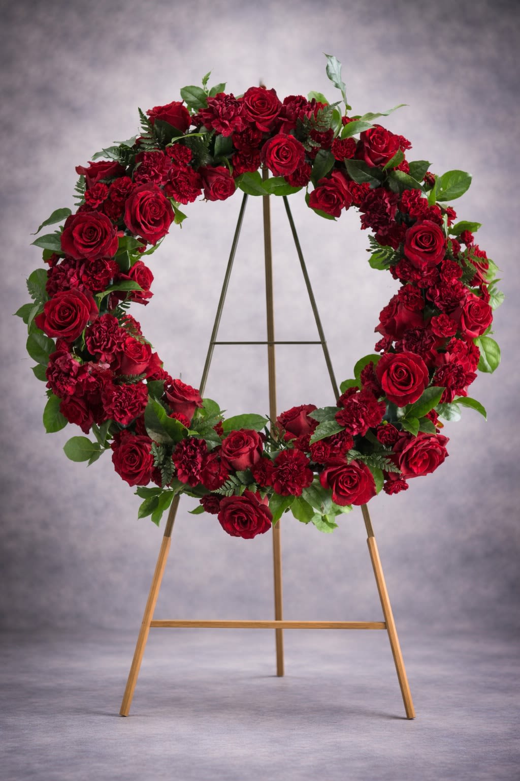 Crimson Remembrance - A striking standing wreath designed with rich red roses and deep red carnations, accented with fresh greenery. This bold and elegant tribute symbolizes love, respect, and lasting remembrance.  Displayed on an easel, this wreath creates a powerful and heartfelt presence at funeral services, memorial gatherings, and celebration-of-life ceremonies.  Substitutions may be necessary based on seasonal availability, but the overall color intensity, fullness, and design integrity will always be maintained.  Delivery available to funeral homes, churches, cemeteries, and residences. Please include service time and location details at checkout.