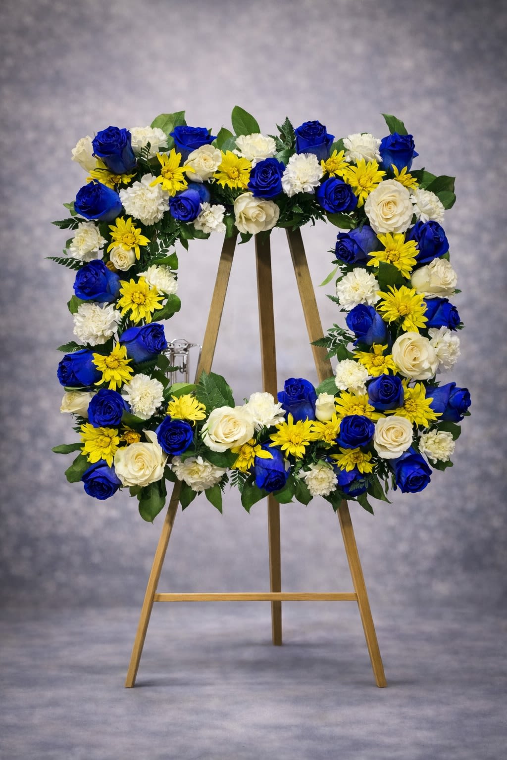 Tribute of Honor - A vibrant standing wreath designed with deep blue roses, white roses, yellow accent blooms, and fresh greenery. This meaningful tribute symbolizes honor, strength, and remembrance.  Displayed on an easel, this arrangement creates a powerful and respectful presence at funeral services, memorials, and celebration-of-life gatherings.  Substitutions may be necessary based on seasonal availability, but the overall color palette, fullness, and wreath shape will always be maintained.  Delivery available to funeral homes, churches, cemeteries, and residences. Please include service time and location details at checkout.